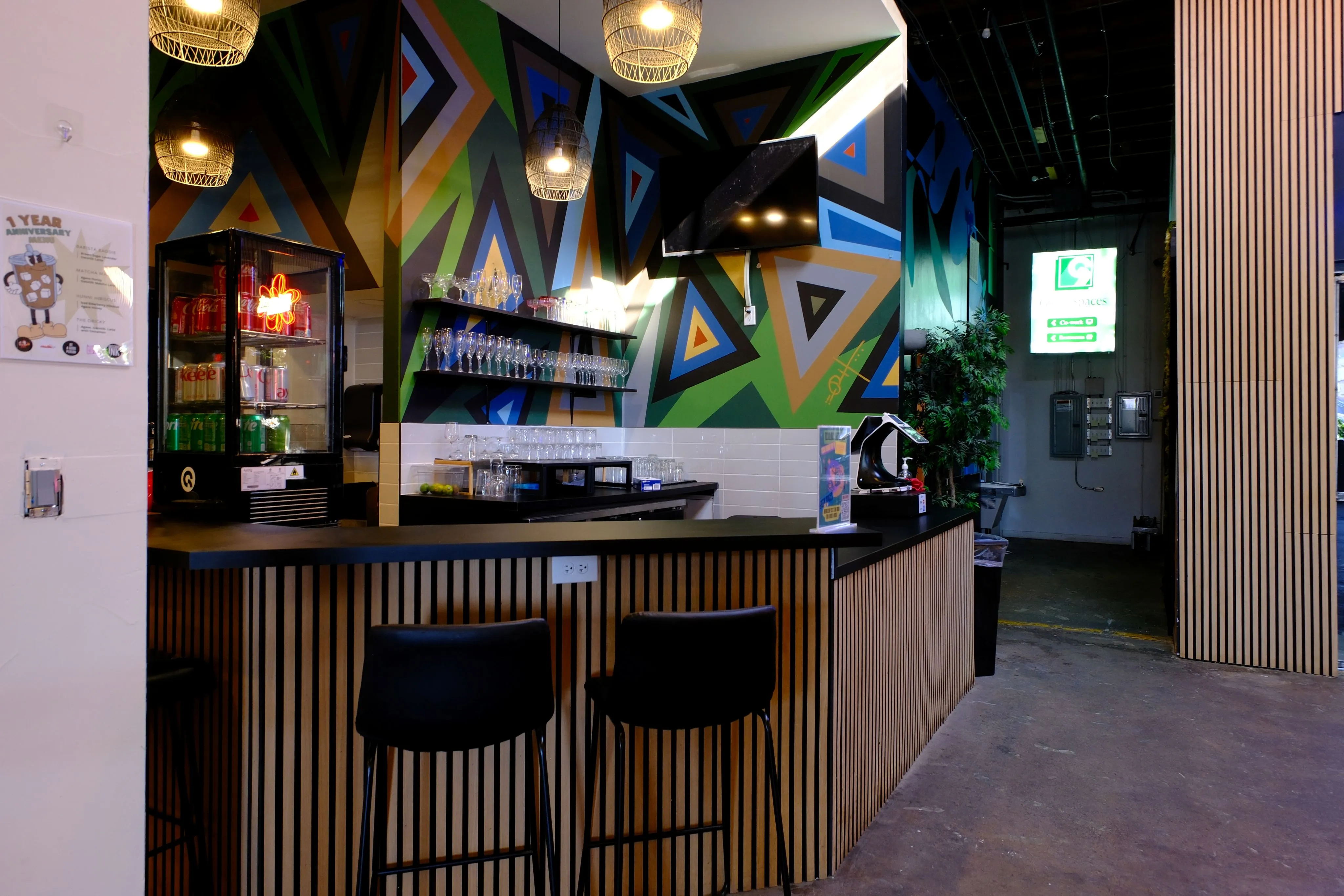 Modern bar area with wooden slat counter, two black stools, colorful geometric wall mural, glassware on shelves, and a small refrigerator with drinks.