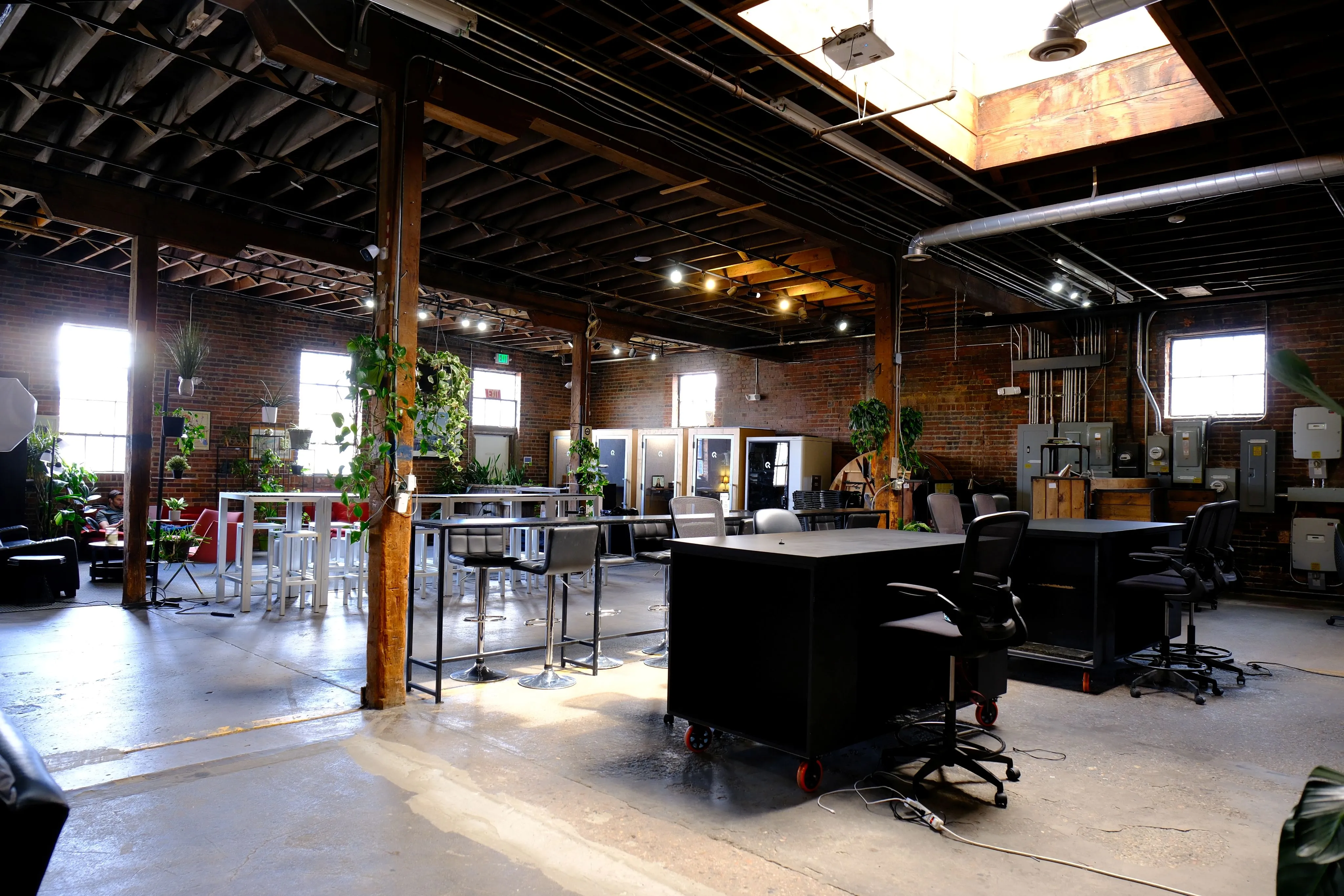 Spacious industrial-style coworking space with wooden beams, high ceilings, lounge chairs, desks, office chairs, and hanging plants.