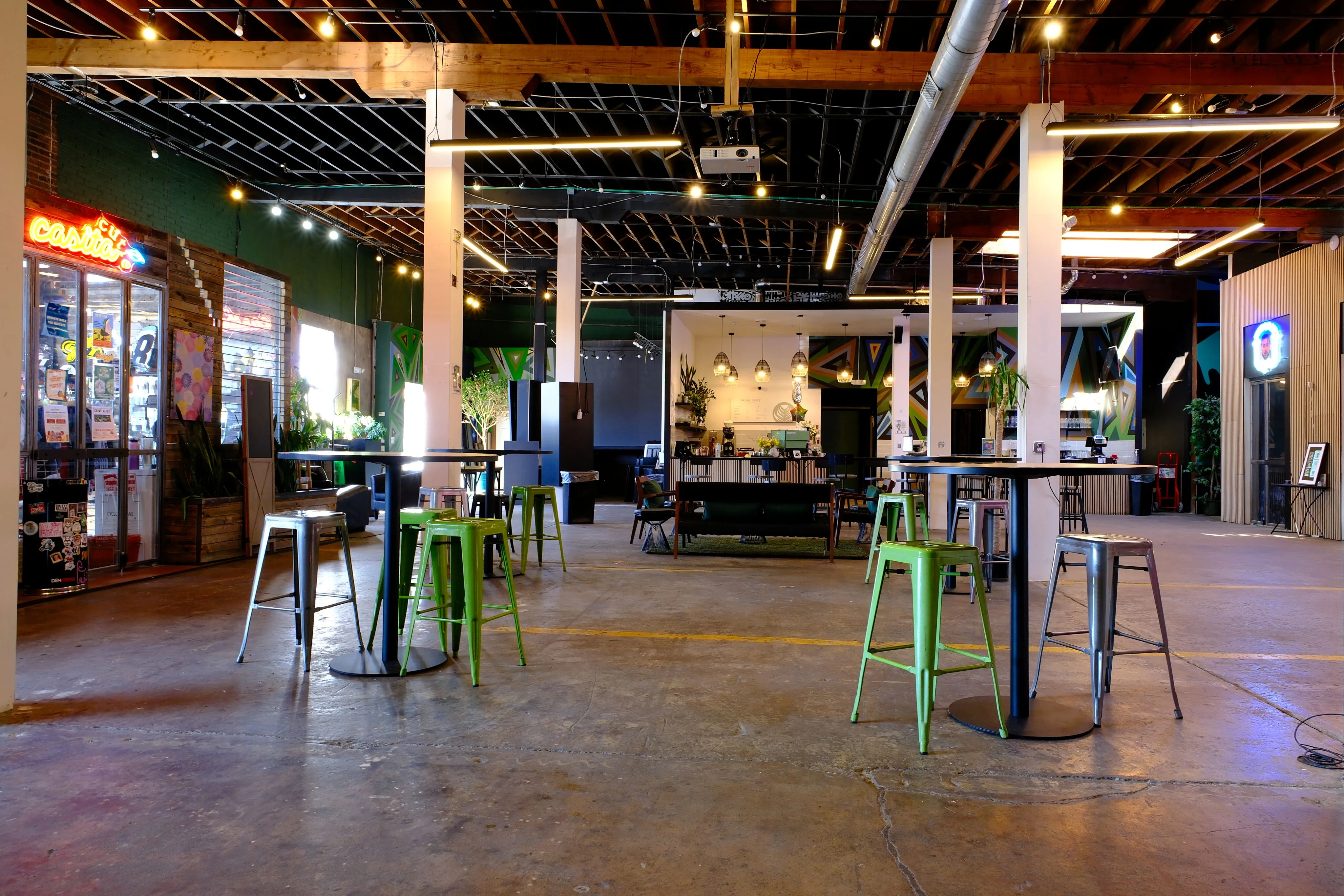 Spacious modern cafe interior with green and silver metal stools around tall tables, exposed wooden ceiling beams, and a counter in the background with hanging lights.