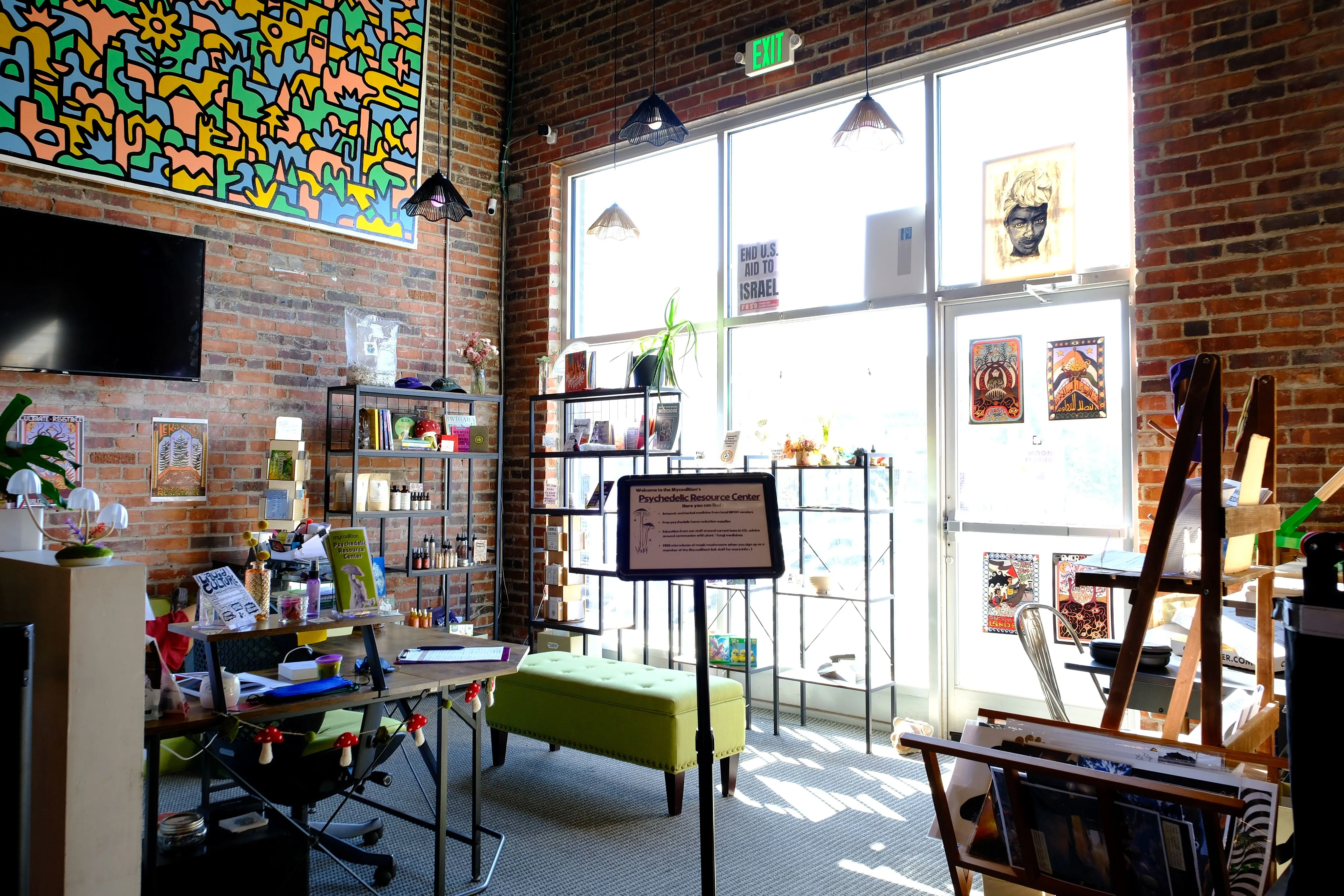 Bright room with brick walls featuring colorful artwork, shelves with various items, a green bench, and a sign for the Mycoalition Psychedelic Resource Center near a glass door with posters.