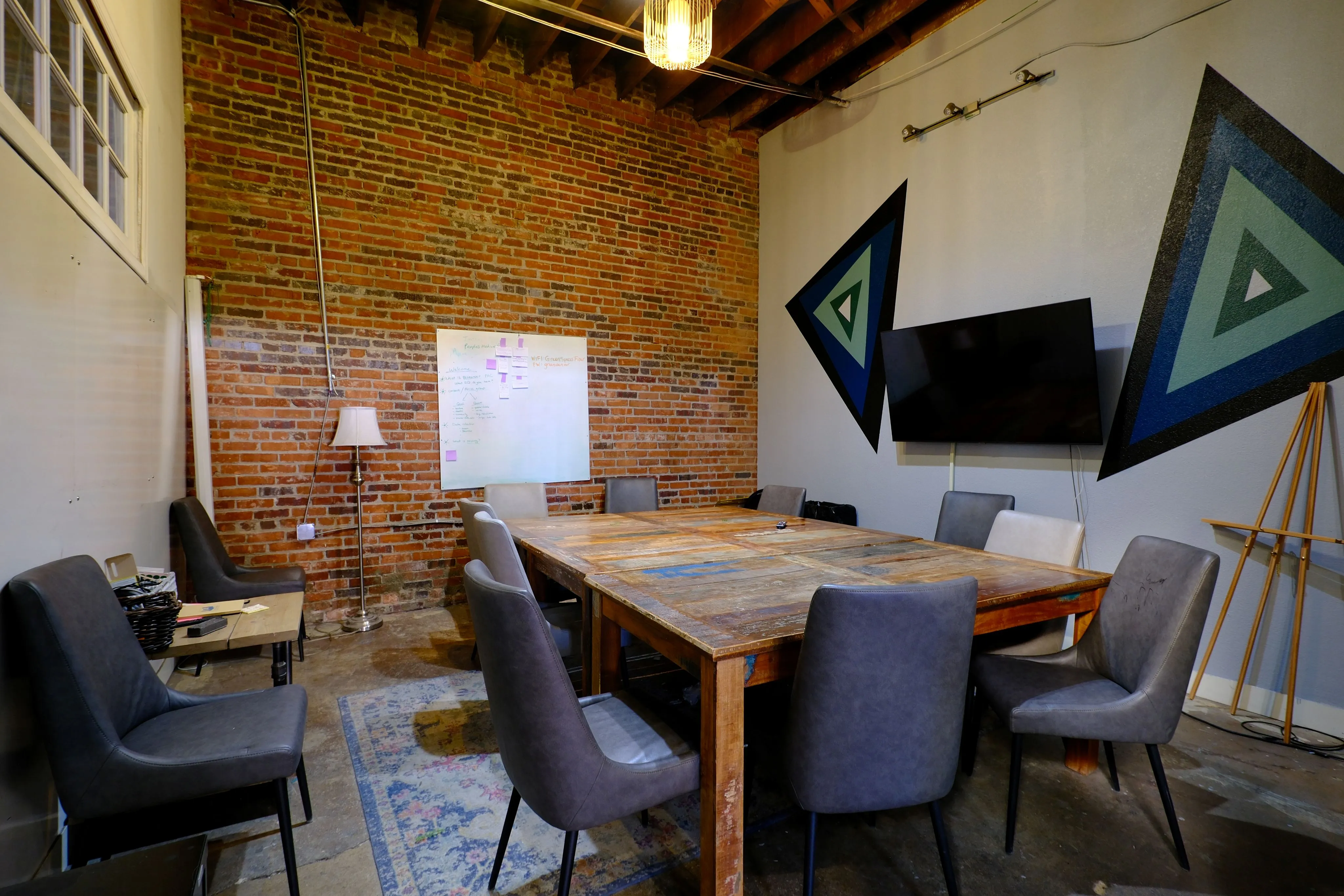 Modern conference room with a large wooden table surrounded by gray chairs, a brick wall, whiteboard, TV screen, and geometric wall art.