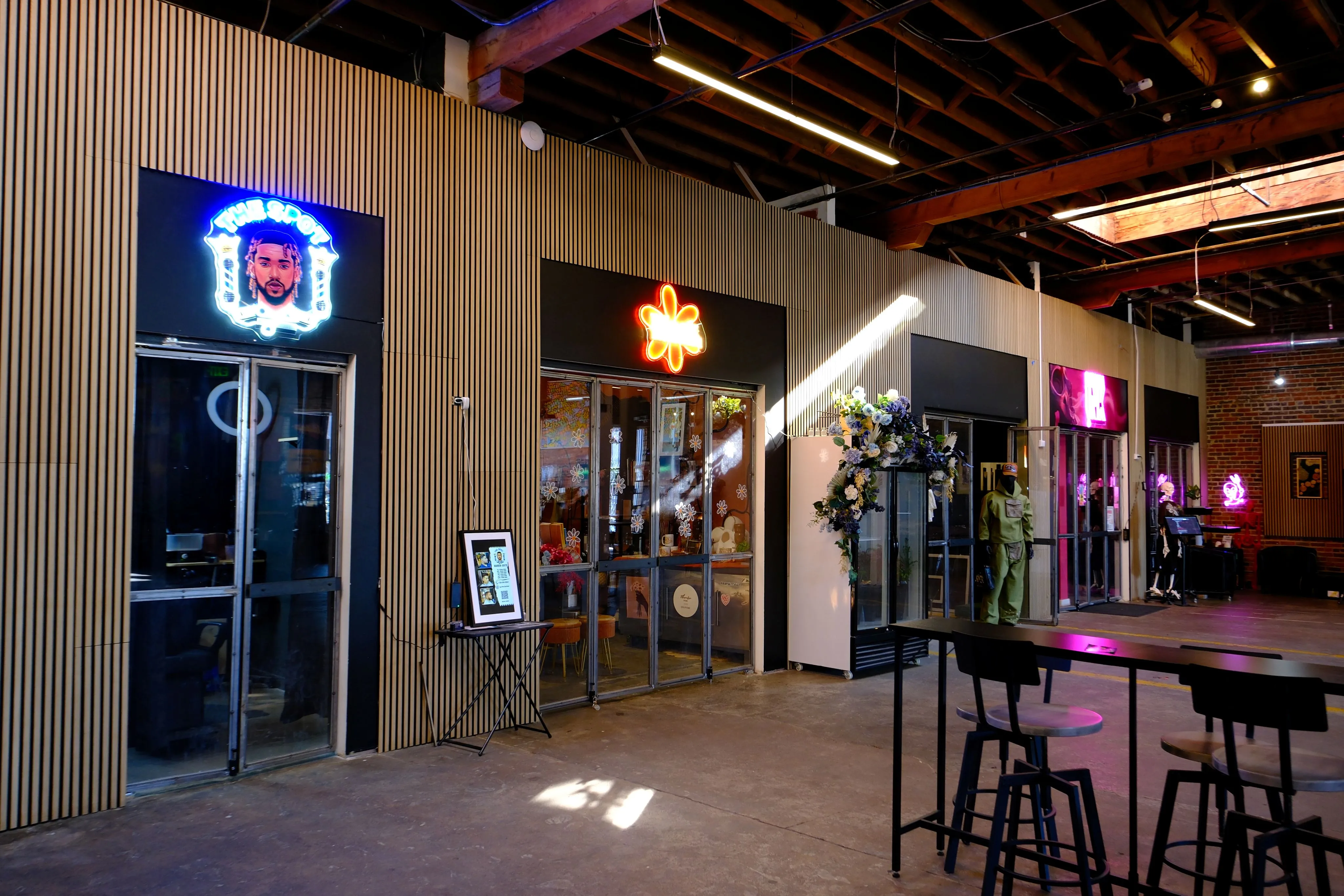 Interior of a modern space with three storefronts featuring neon signs, wooden slat walls, and a seating area with a tall table and chairs.