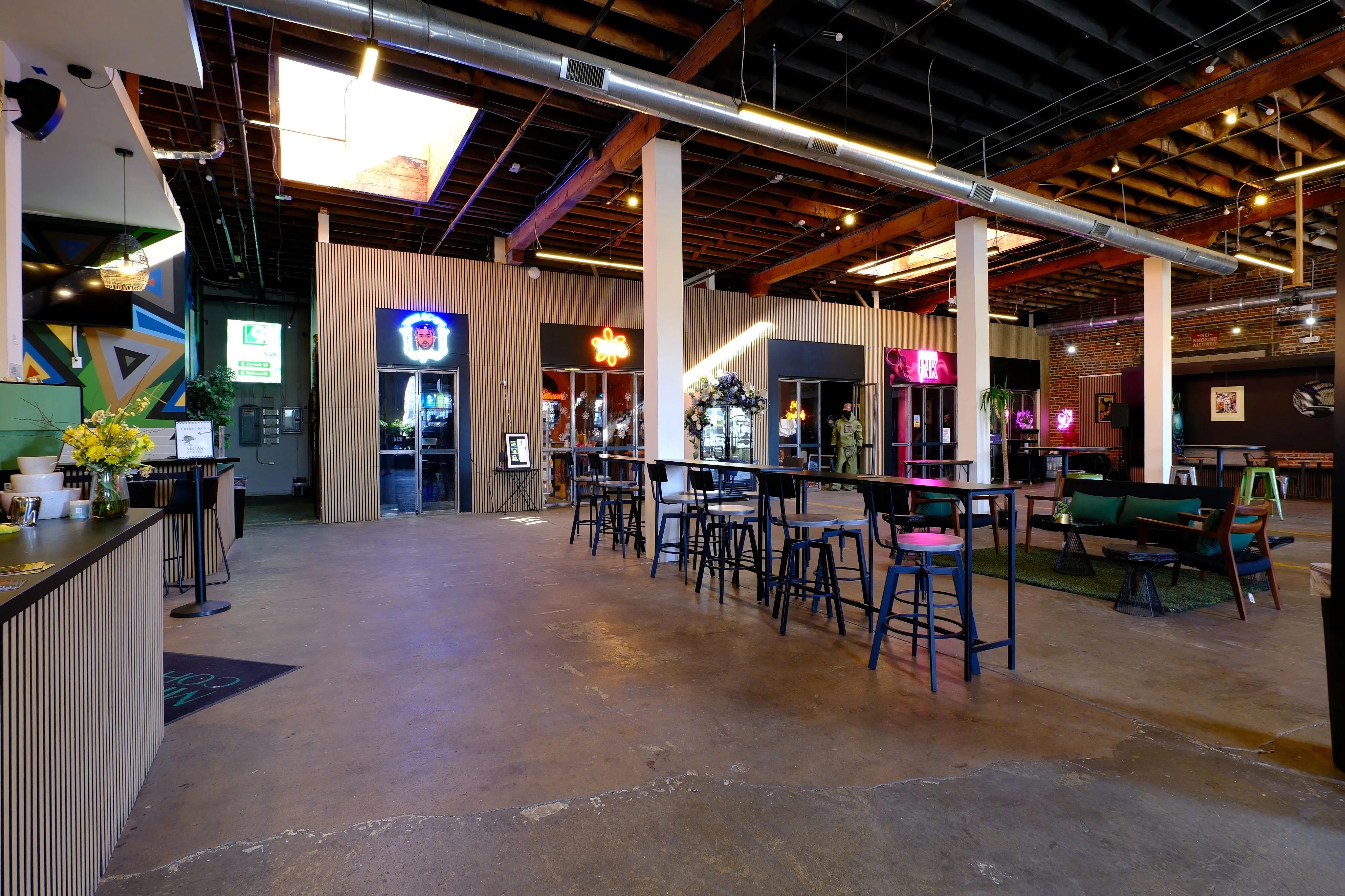 Spacious modern indoor venue with high wooden ceilings, tall stools at long tables, neon signs, and lounge seating.