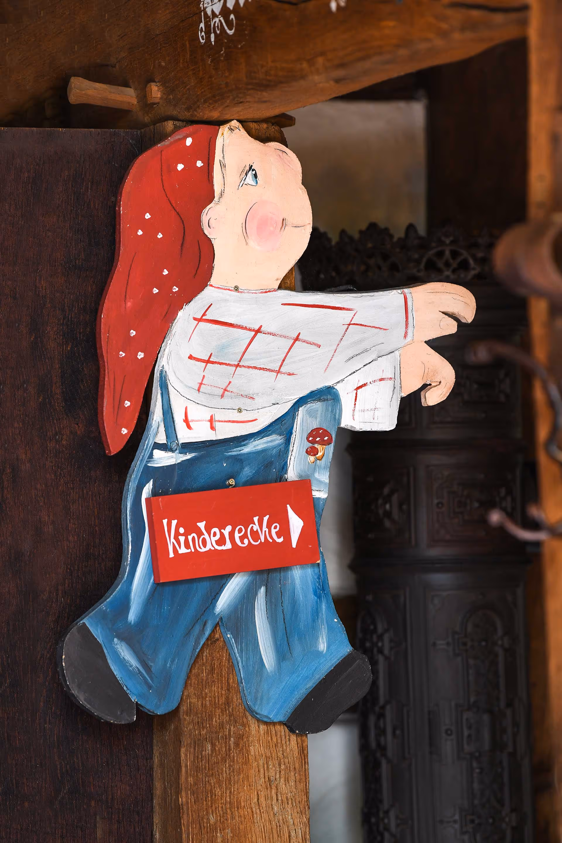 Ein kleiner Mensch mit Latzhose und einer Mütze mit einem Schild "Kinderecke" um dem Hals.