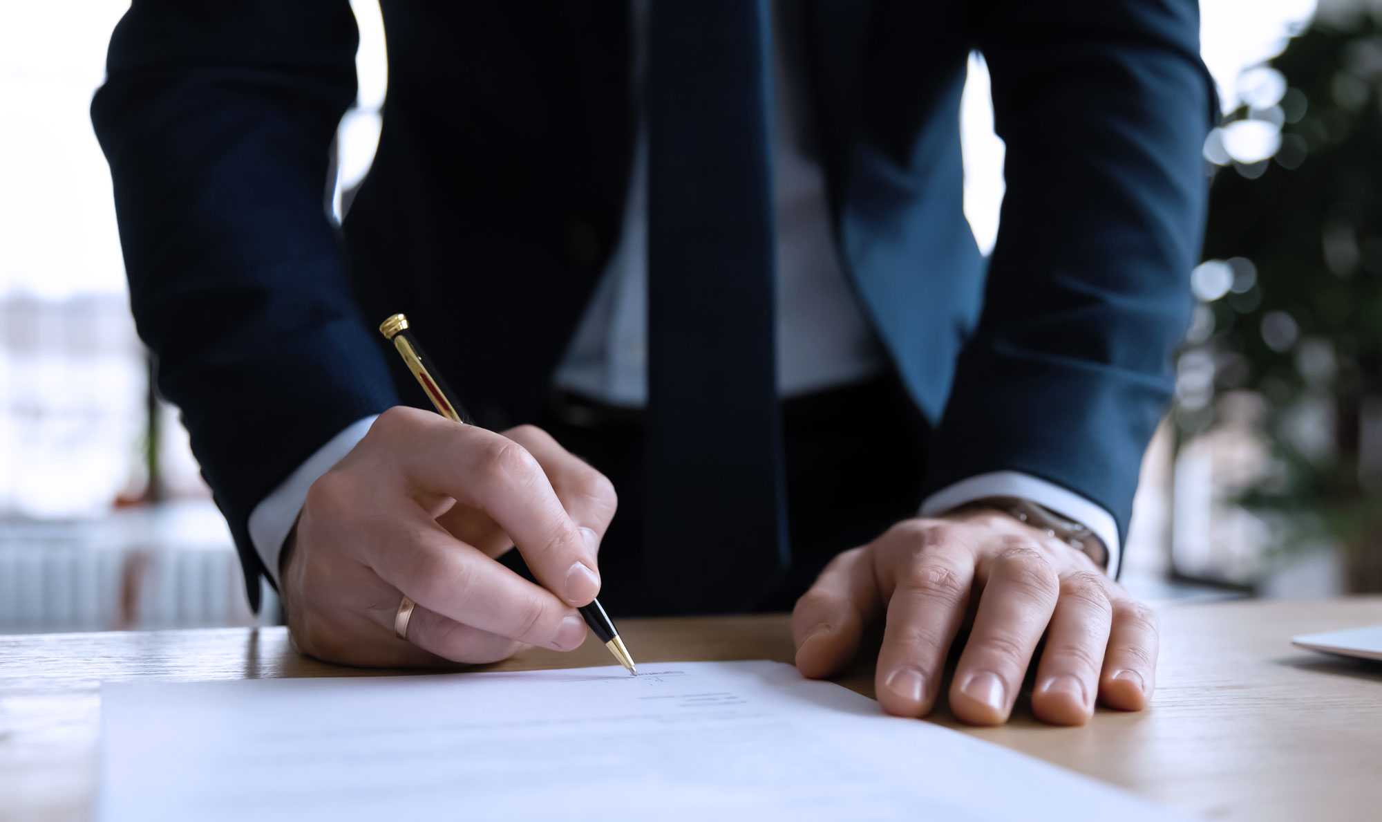 Man in a suit signing a document on a wooden table with a black and gold pen.