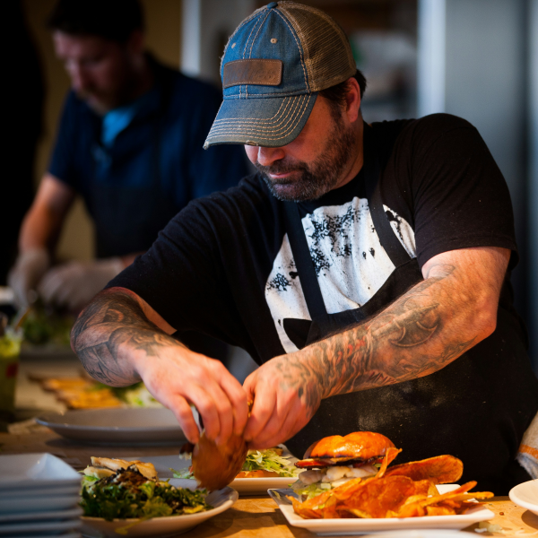 Un cuisinier tatoué portant une casquette bleue prépare des plats dans une cuisine, ajoutant des ingrédients sur une assiette avec des tacos et une salade.