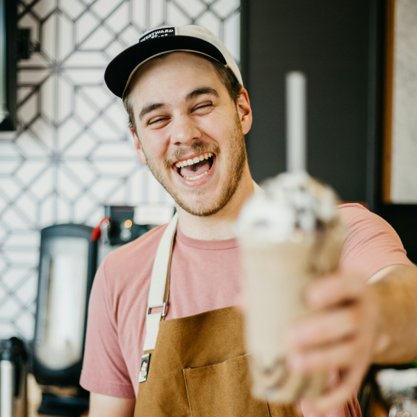Jeune homme souriant portant une casquette et un tablier, tenant un milkshake avec chantilly et une paille à la main.