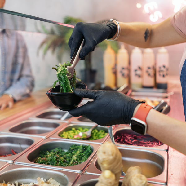 Une personne portant des gants noirs utilise une pince pour servir une salade d'algues dans un petit bol noir dans un comptoir de self-service avec divers ingrédients.