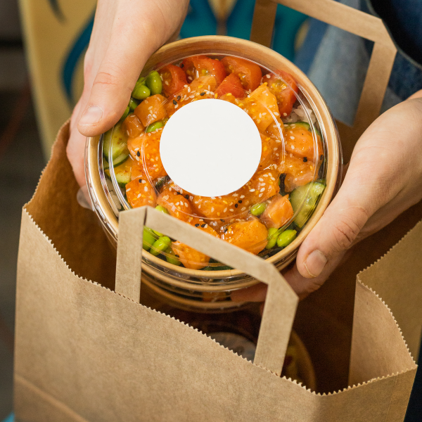 Deux mains tenant un bol transparent avec une salade composée de morceaux de saumon, tomates cerises, edamame et graines, placé dans un sac en papier.