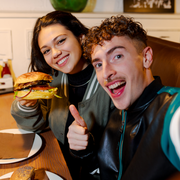 Deux jeunes souriants, une femme tenant un hamburger et un homme montrant un pouce levé dans un restaurant.