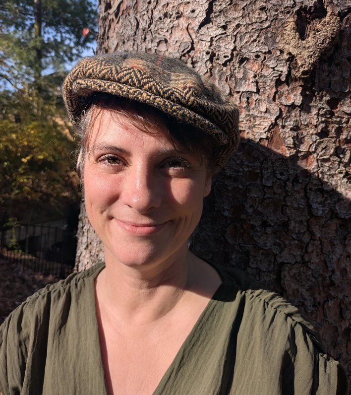 Smiling person wearing a patterned flat cap and olive green shirt standing in front of a large tree trunk outdoors.