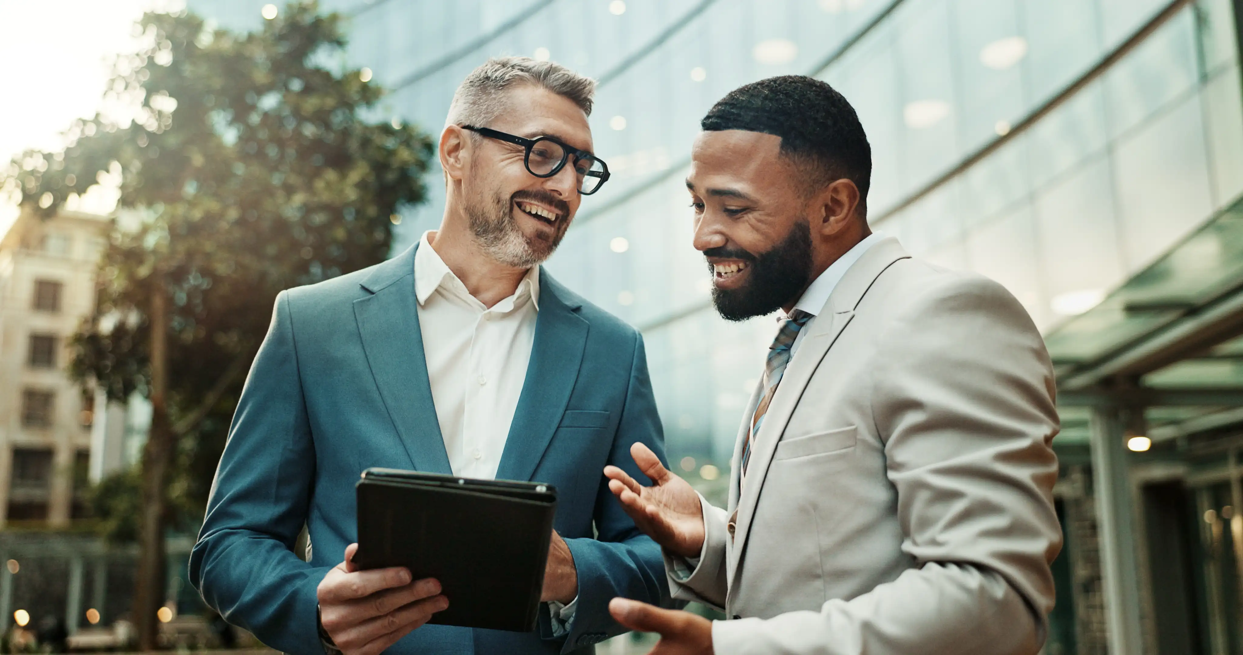 Zwei Geschäftsmänner lächeln und unterhalten sich draußen, während sie ein Tablet betrachten.