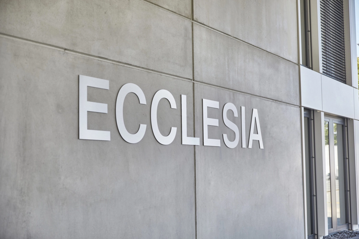 Large white letters spelling 'ECCLESIA' mounted on a smooth gray concrete wall outside a modern building.