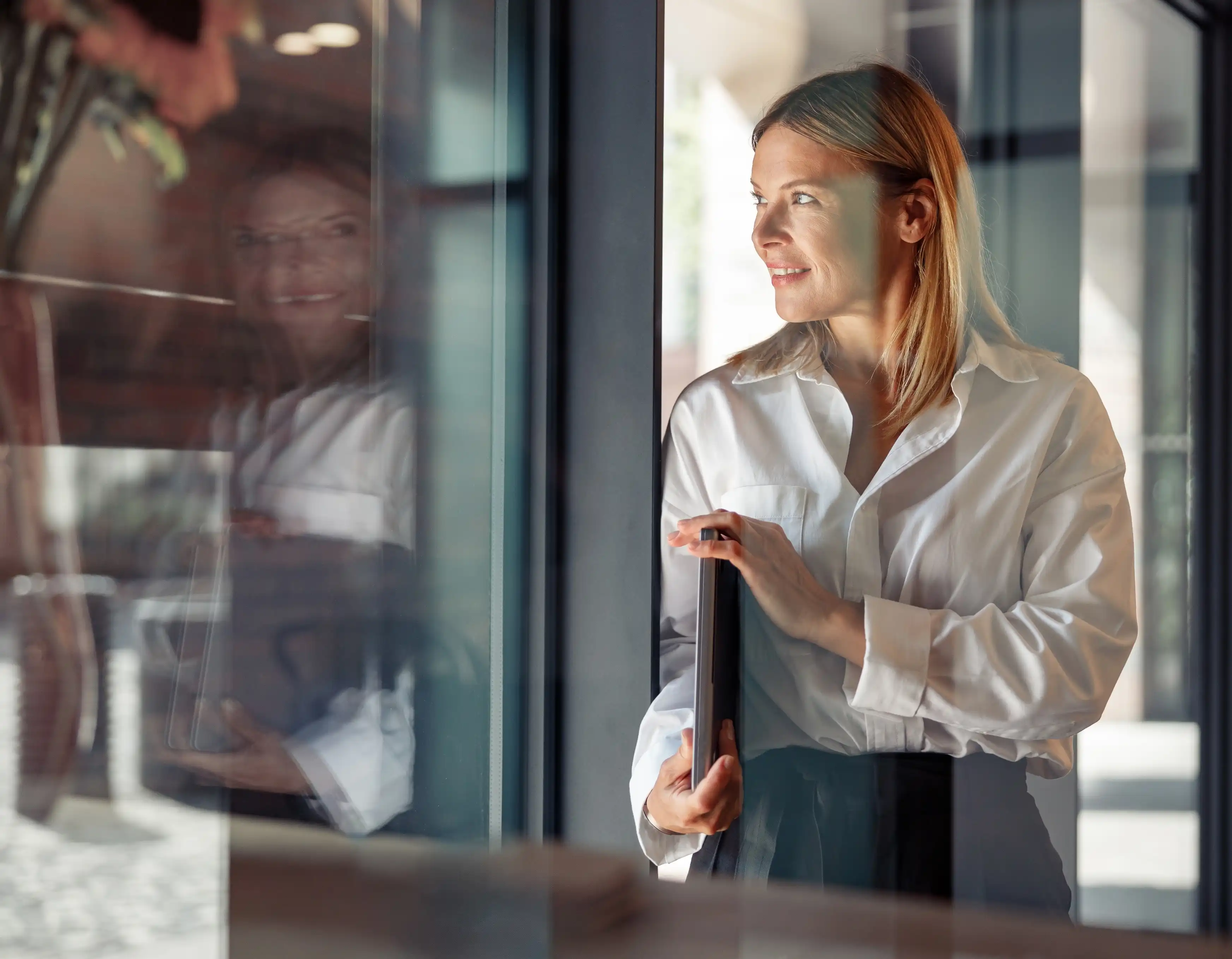 Smiling woman in a white shirt holding a laptop and looking out a window with her reflection visible.