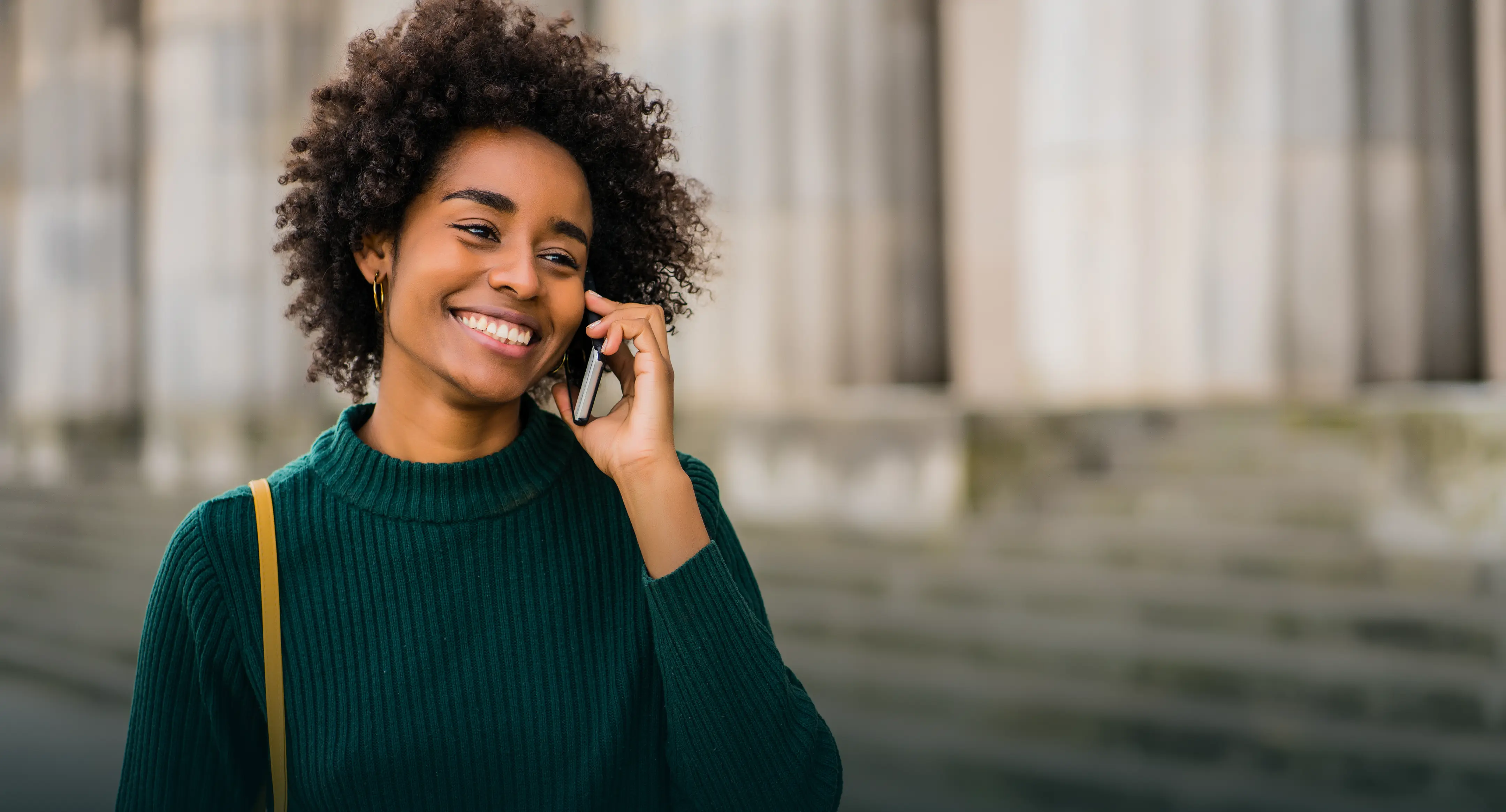 Frau mit lockigem Haar, die draußen lächelnd telefoniert.