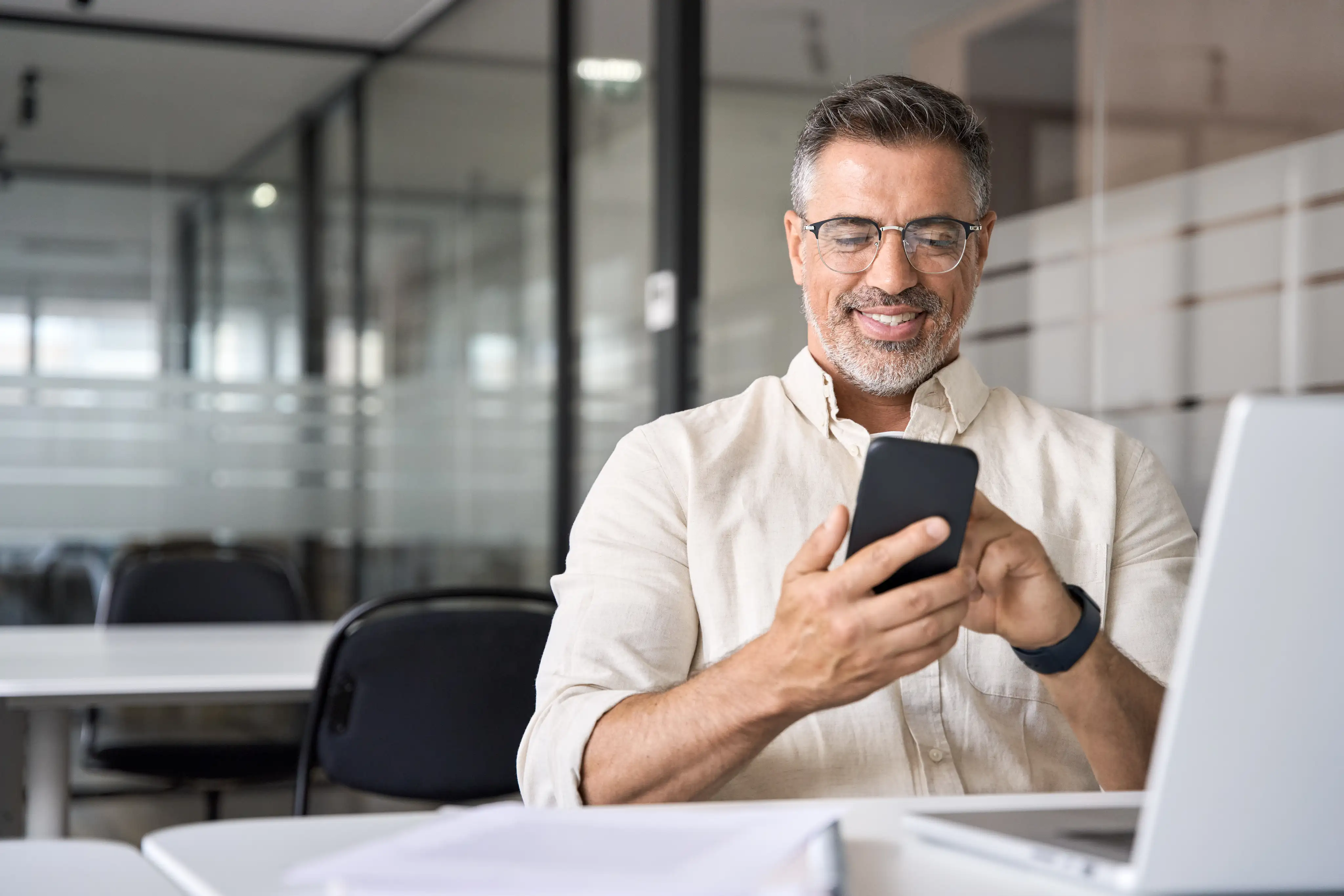 Mann mit Brille und grauem Bart lächelt und benutzt ein Smartphone in einem modernen Büro.