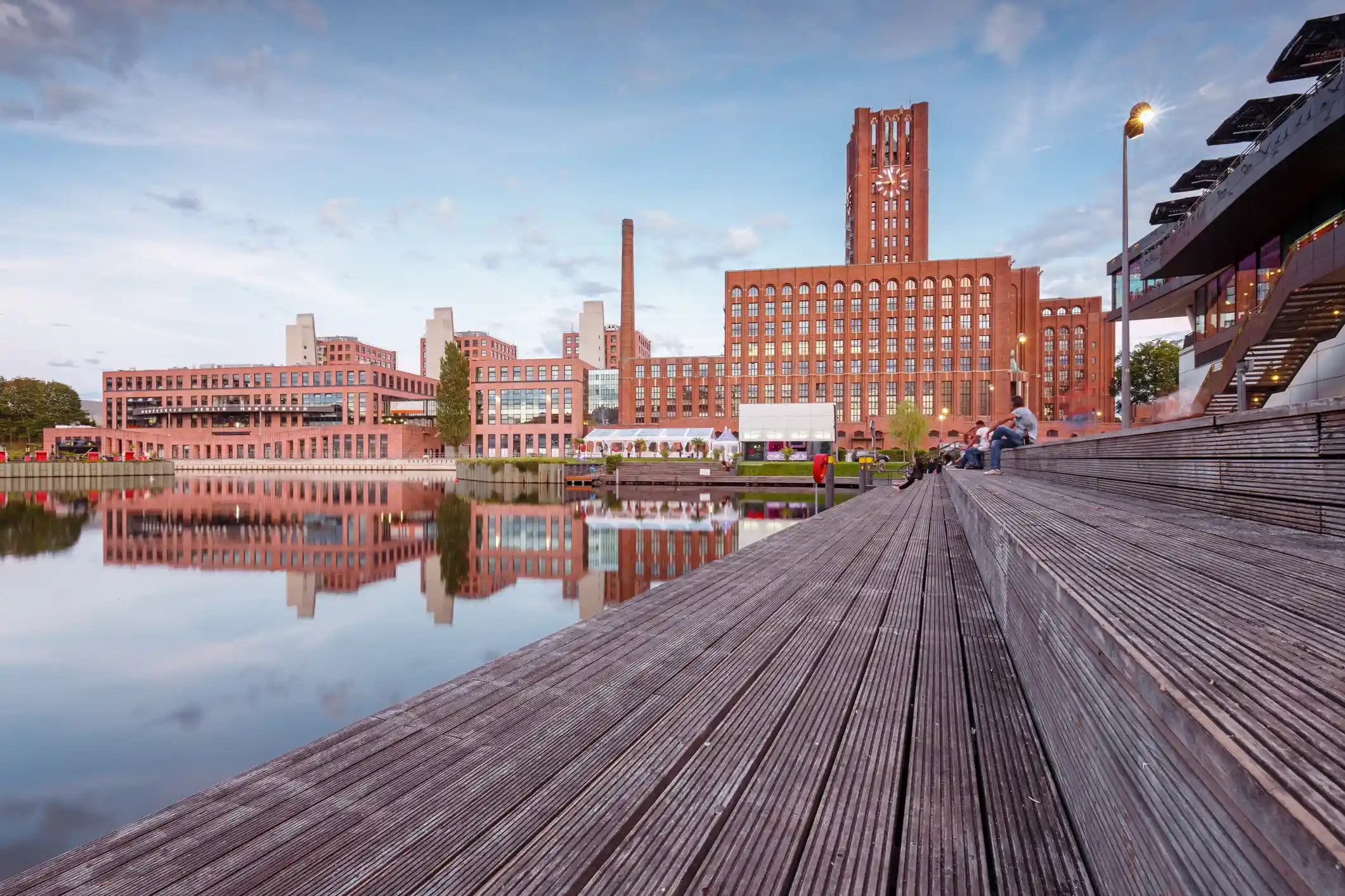 Großer Backsteingebäudekomplex mit Uhrenturm spiegelt sich in ruhigem Wasser neben hölzernen Stufen, auf denen einige Menschen sitzen.