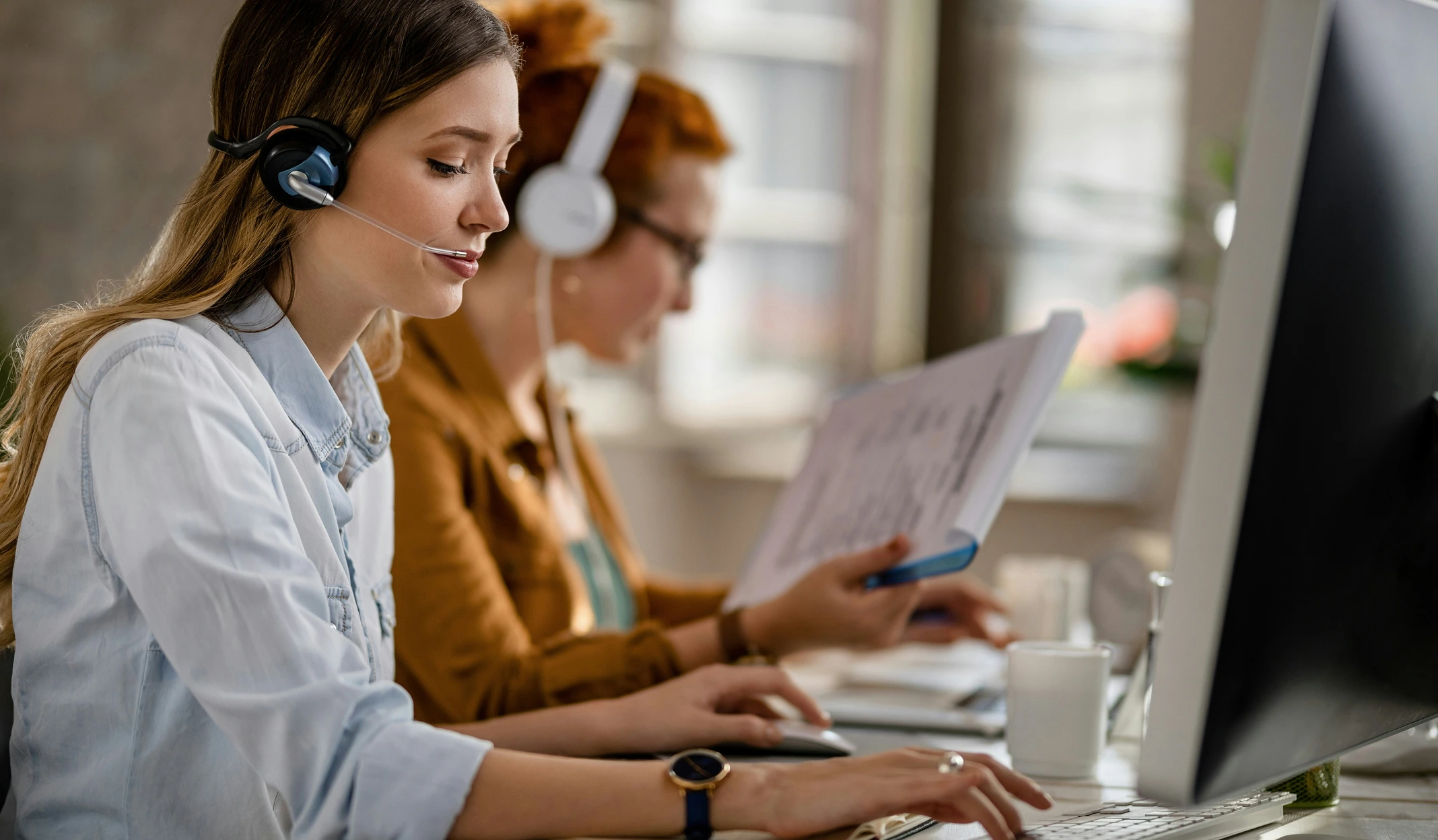 Zwei Frauen tragen Headsets und arbeiten am Computer, eine tippt auf der Tastatur, die andere liest Dokumente.