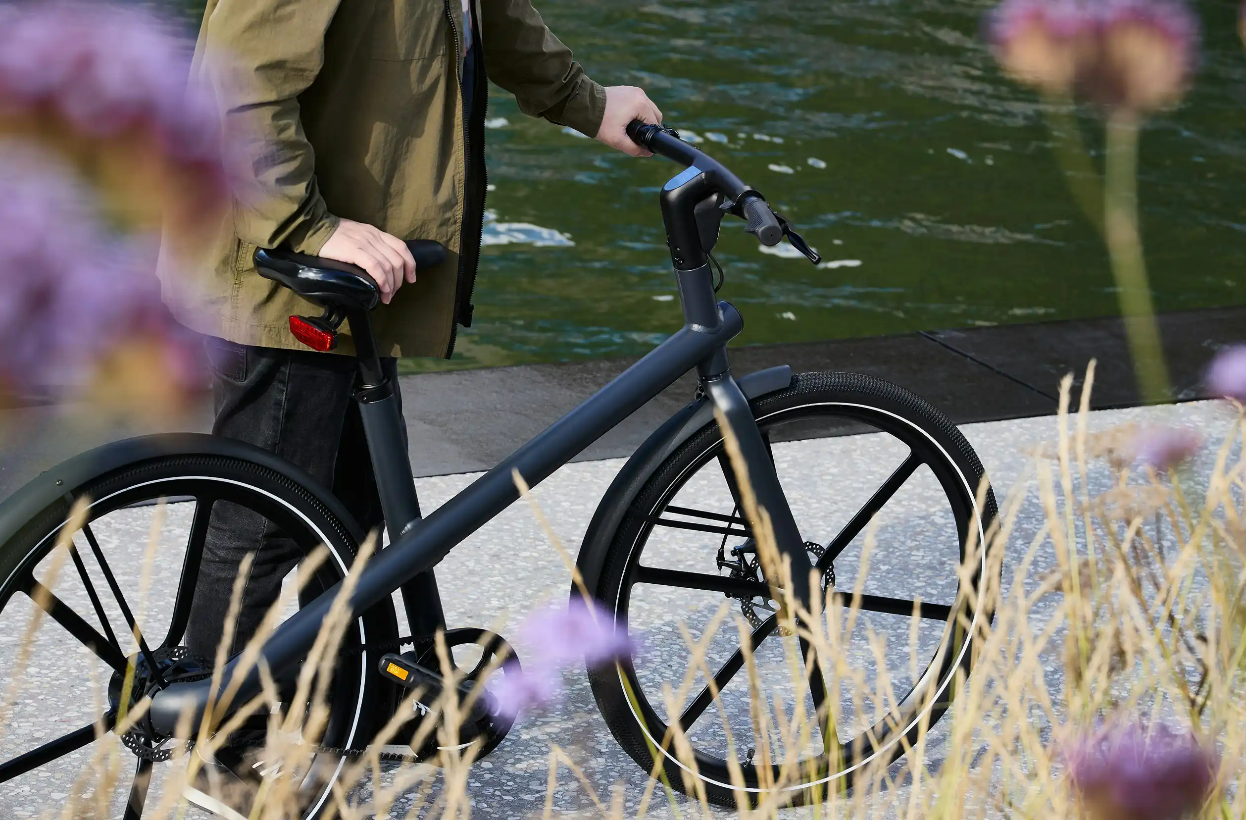 Person mit grüner Jacke steht neben einem schwarzen Fahrrad am Ufer eines Flusses mit Blumen im Vordergrund.