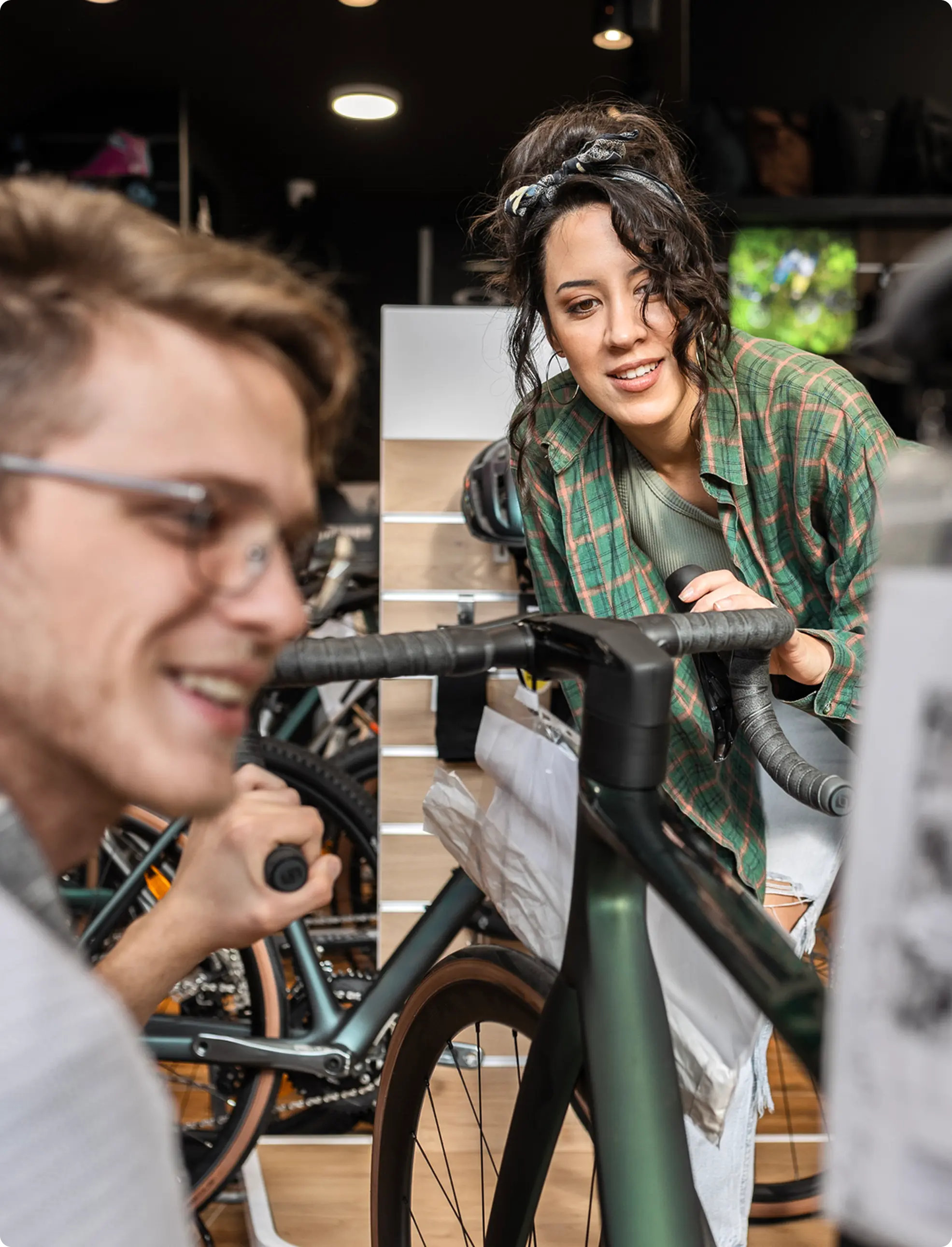 Eine Frau in kariertem Hemd beugt sich lächelnd über den Lenker eines Fahrrads, während ein Mann in Brille daneben steht.