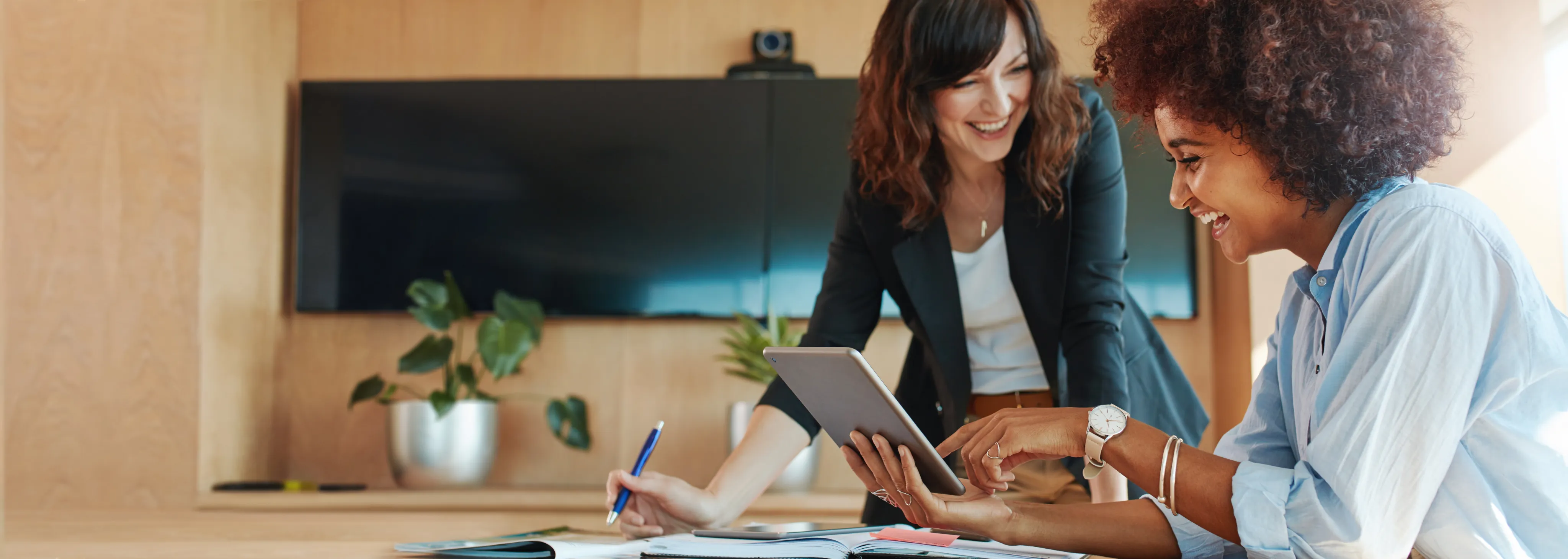Zwei Frauen arbeiten lachend zusammen mit einem Tablet und Notizen in einem modernen Büro.