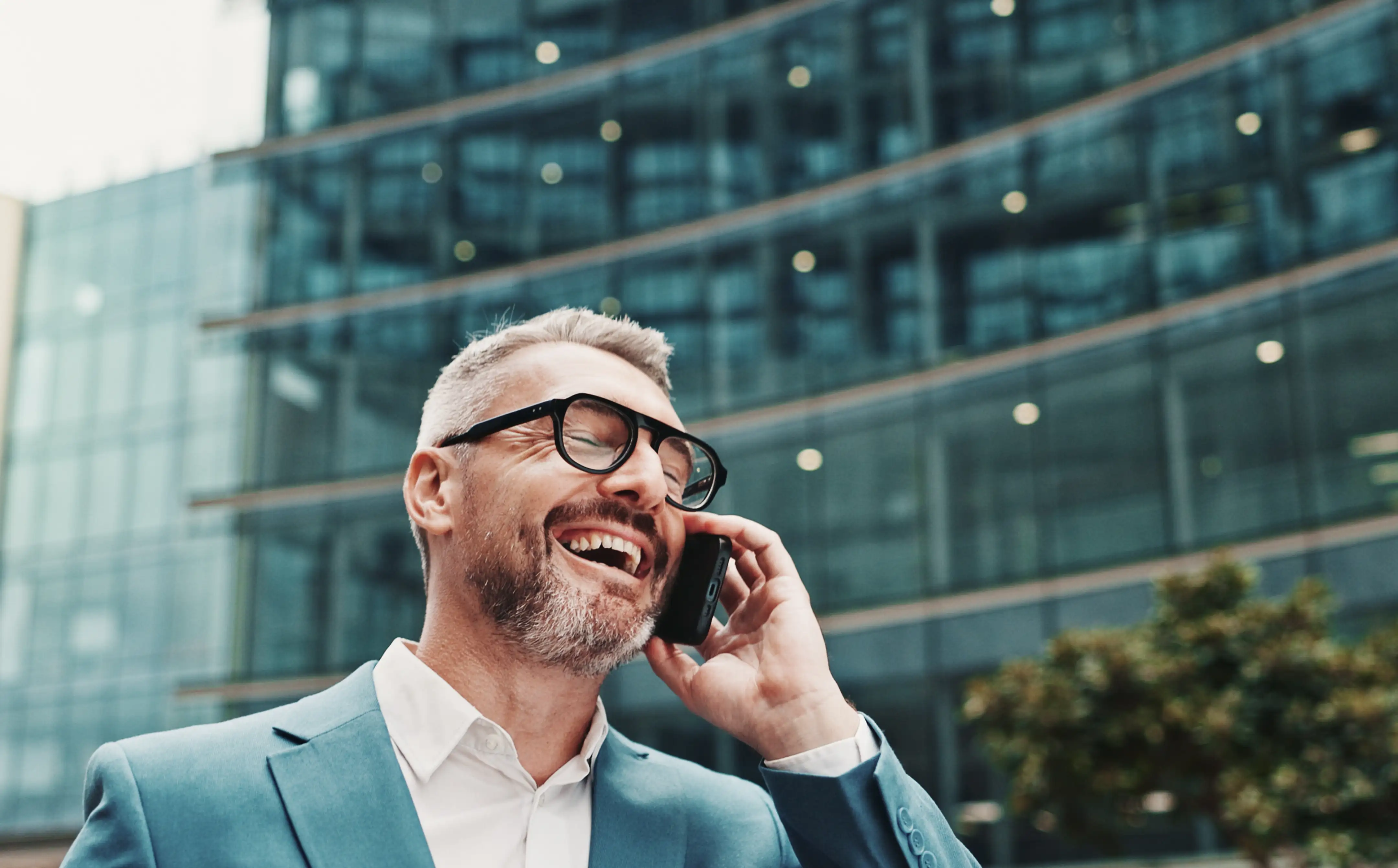 Glücklicher Geschäftsmann mit Brille, der draußen vor einem modernen Glasgebäude telefoniert.