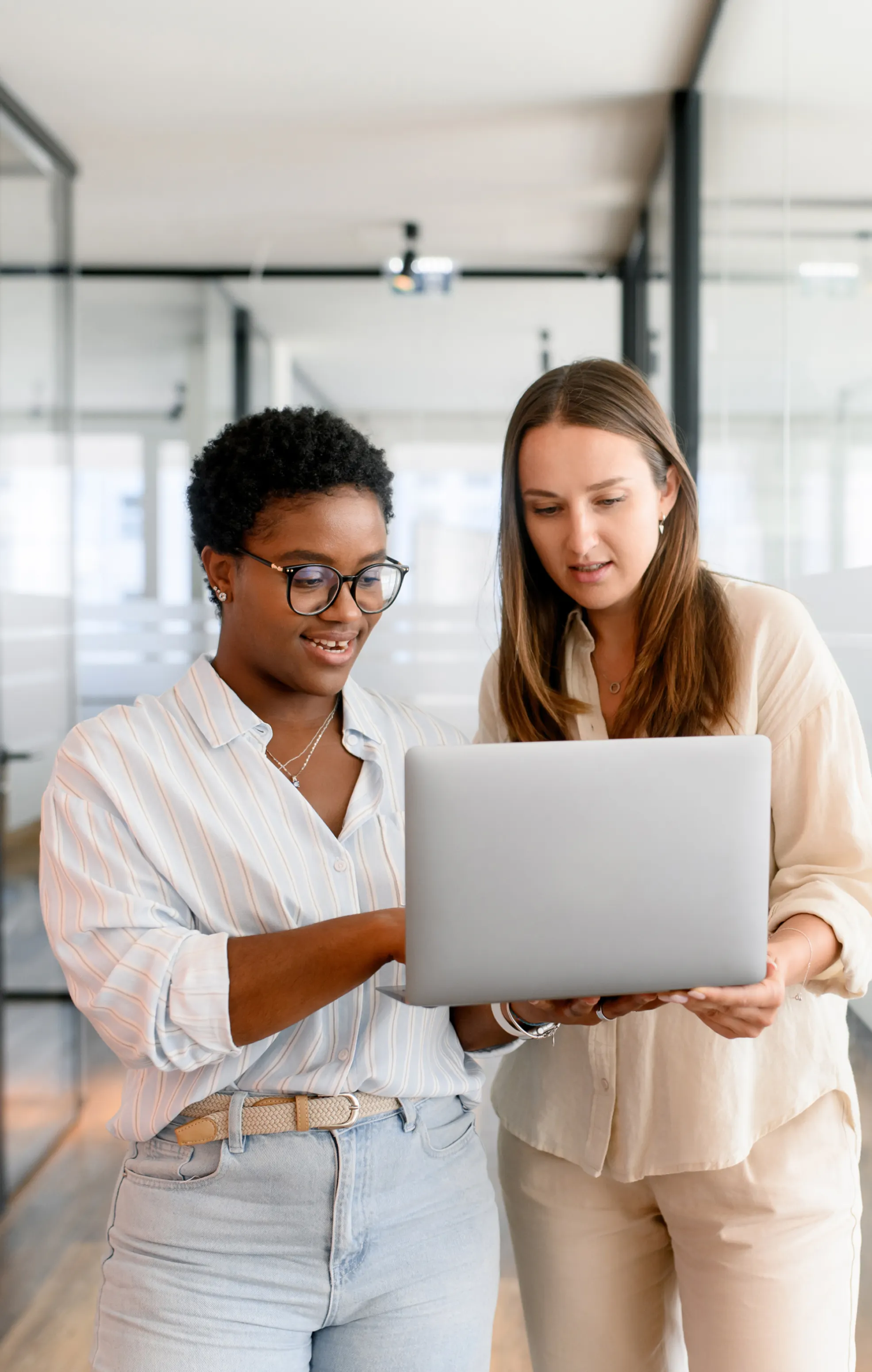 Zwei Frauen stehen in einem modernen Büro und betrachten gemeinsam einen geöffneten Laptop.
