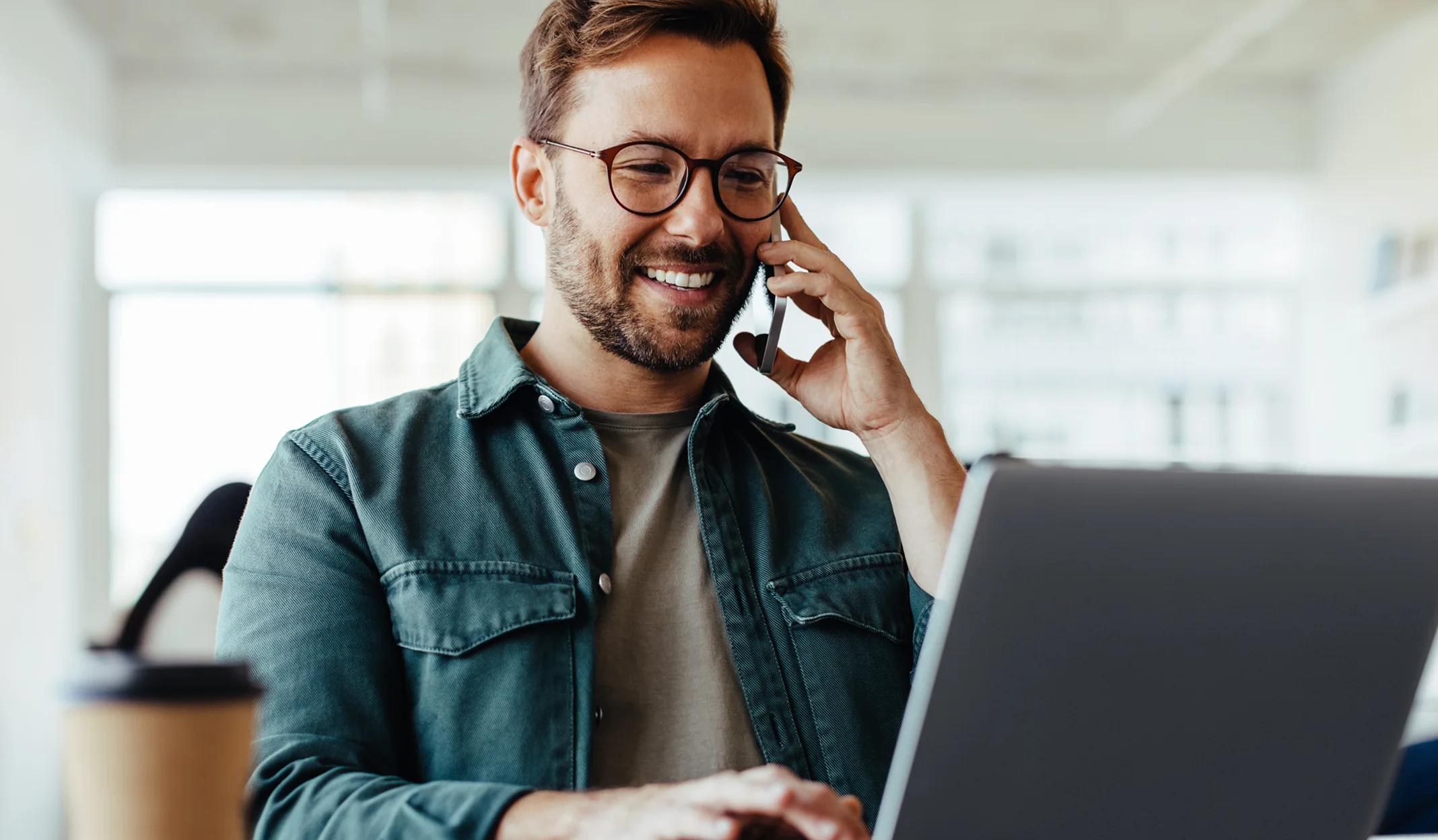 Lächelnder Mann mit Brille telefoniert und arbeitet an einem Laptop in einem hellen Büro.