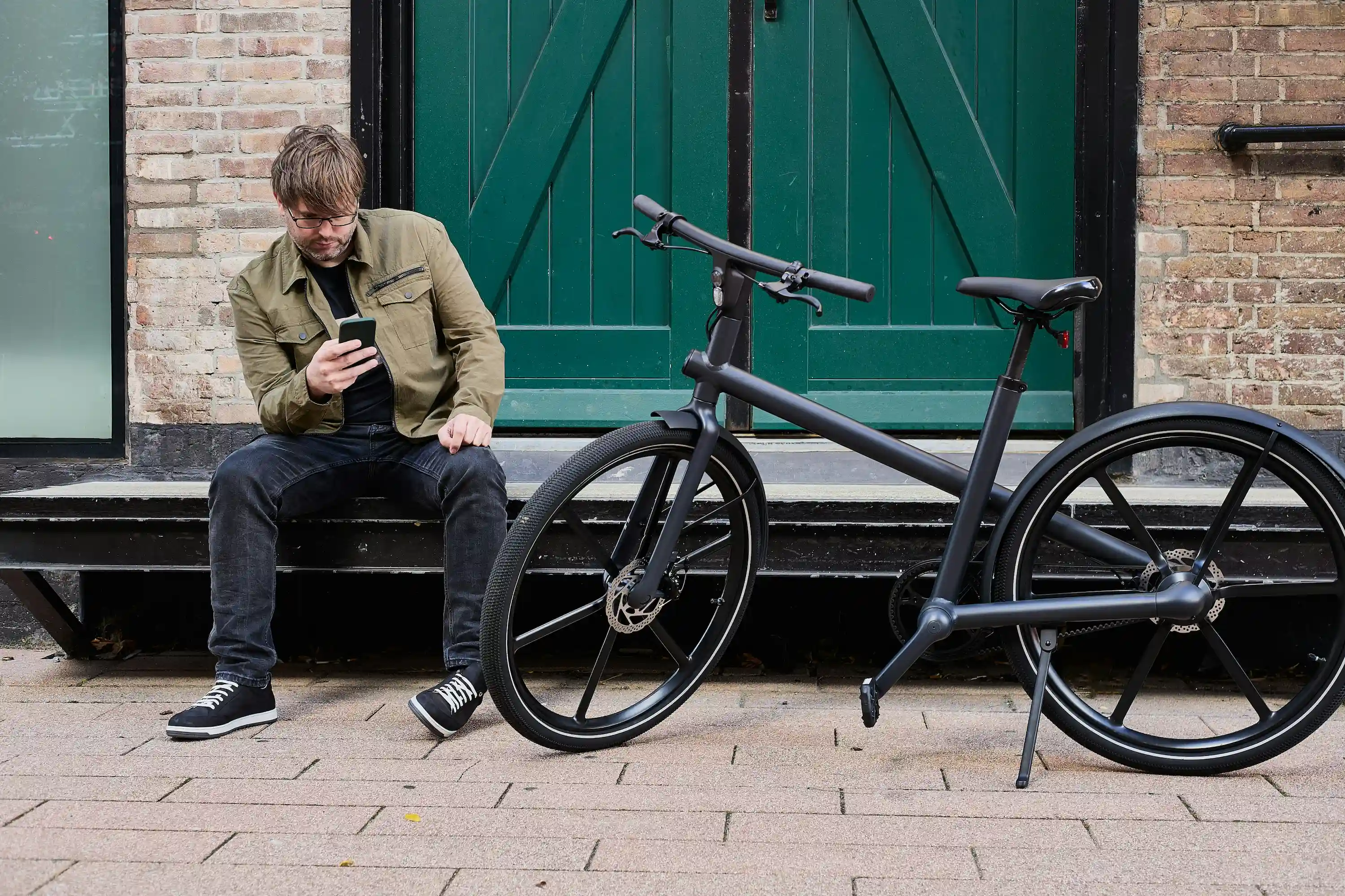 Ein Mann mit Brille und olivgrüner Jacke sitzt auf einer Bank und schaut auf sein Handy, neben ihm steht ein modernes schwarzes Fahrrad.