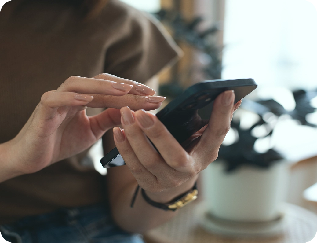 Jemand hält ein Smartphone und bedient es mit der anderen Hand, im Hintergrund unscharfe Pflanzen.