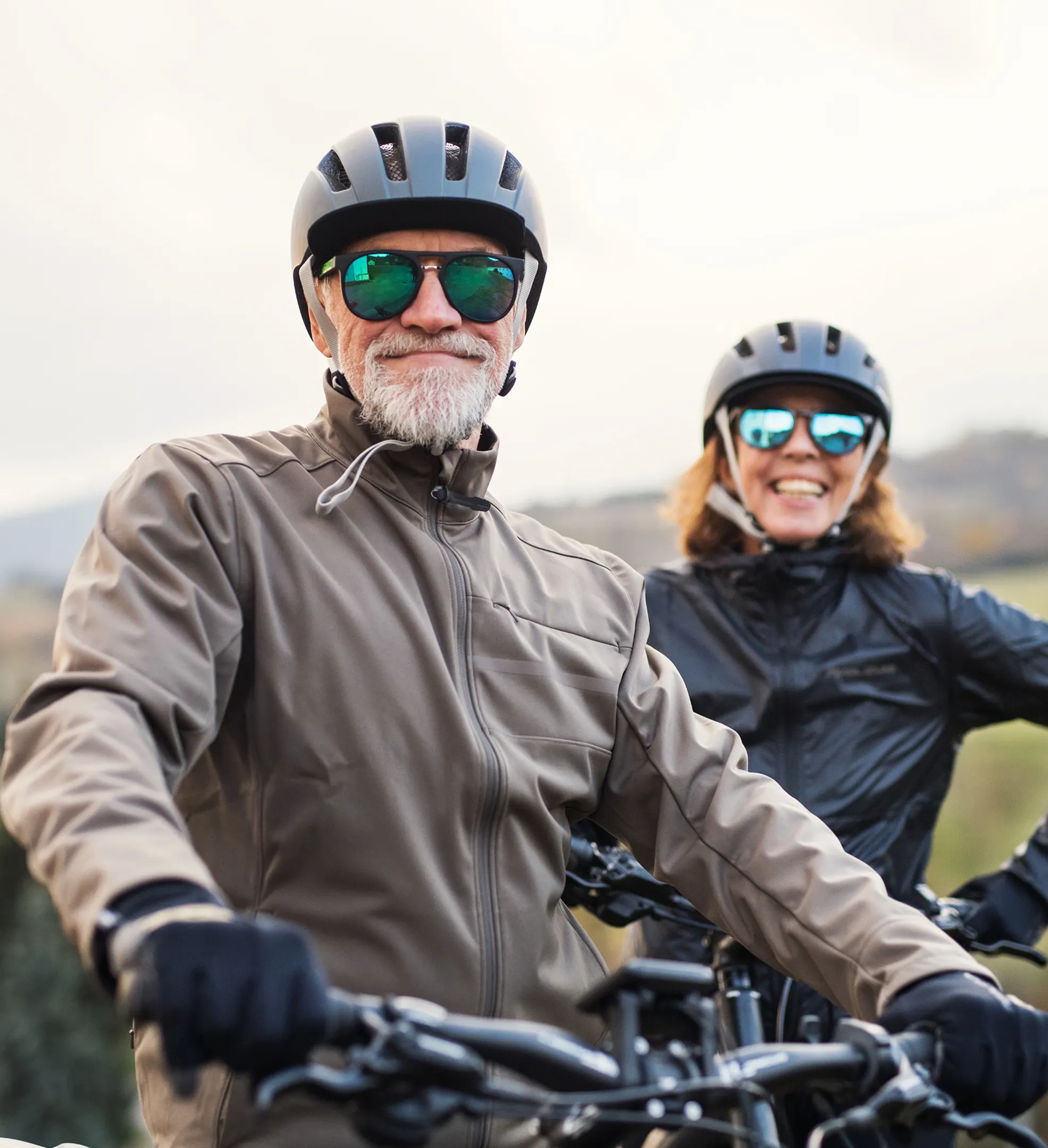 Älteres Paar mit Fahrradhelmen und Sonnenbrillen lächelt und posiert draußen für ein Foto.