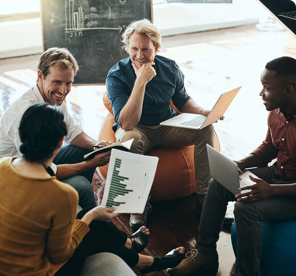 Vier Personen in einem informellen Meeting, die mit Laptops und Diagrammen zusammenarbeiten und lächeln.