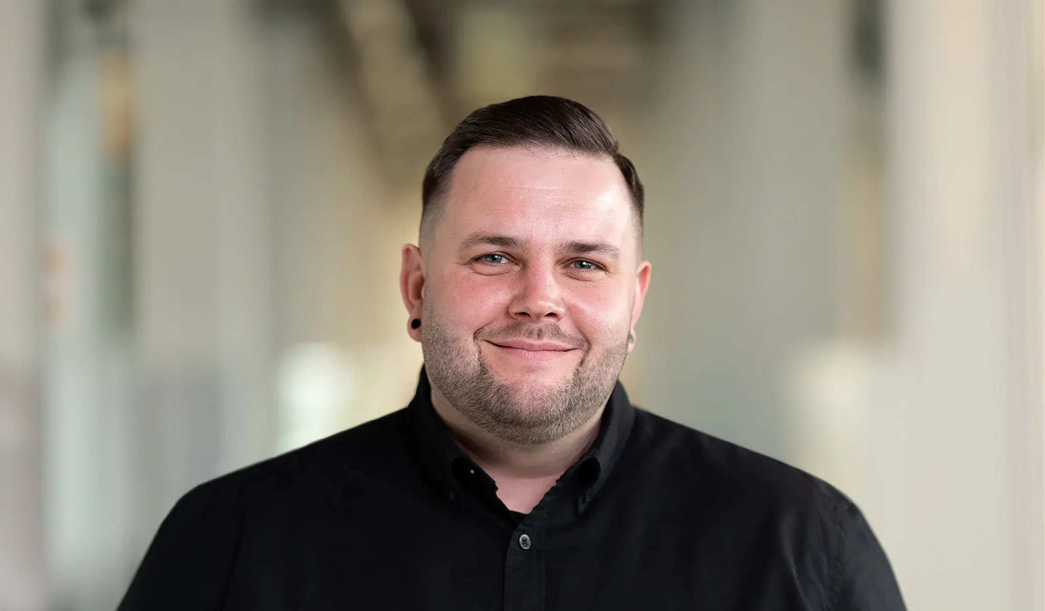 Porträt eines lächelnden Mannes mit kurzem dunklem Haar und Bart, der ein schwarzes Hemd trägt.