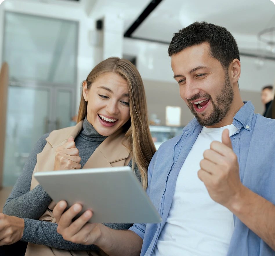 Junge Frau und Mann schauen lächelnd auf ein Tablet und zeigen eine freudige Reaktion.