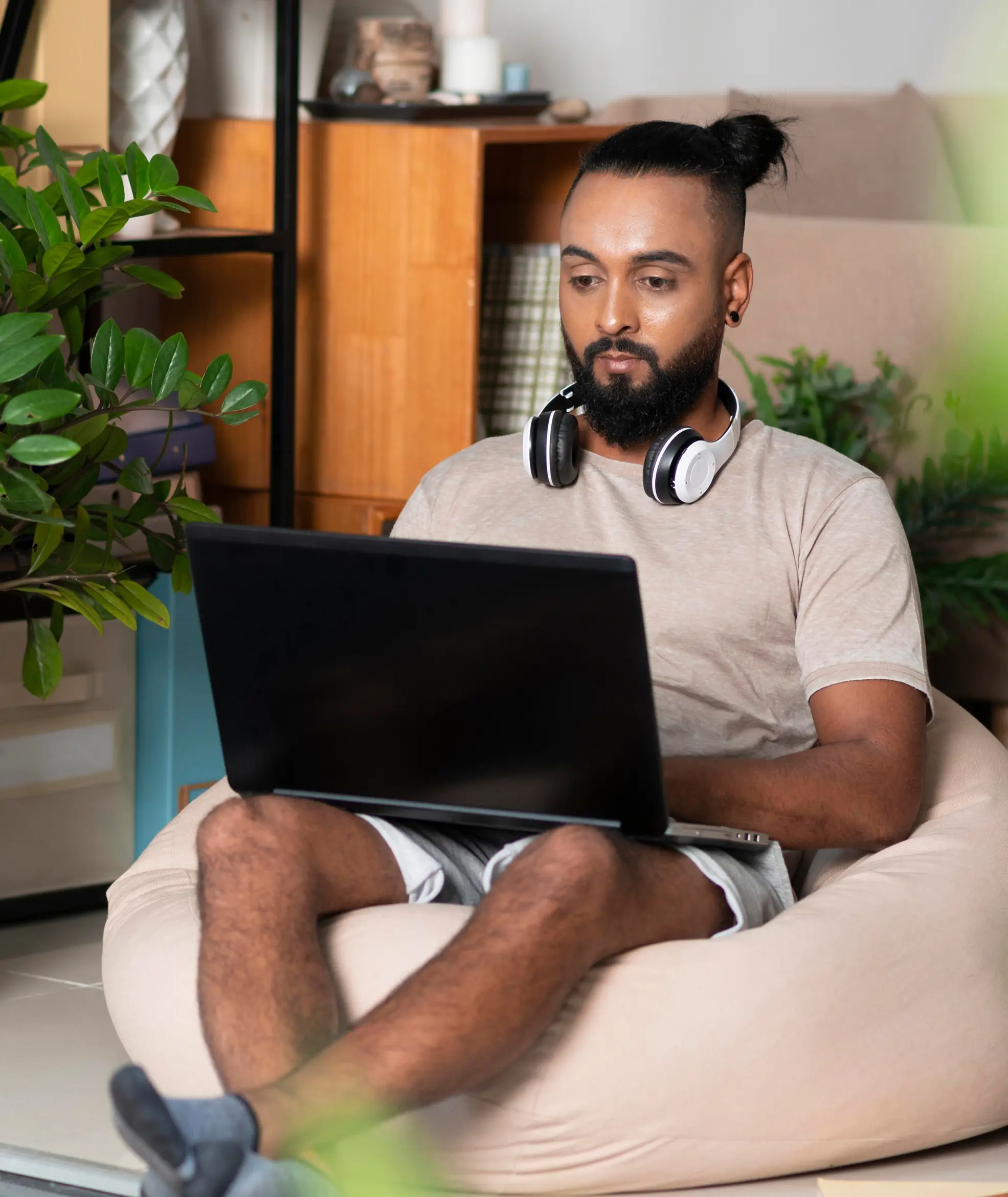 Mann mit Zopf und Kopfhörern um den Hals sitzt entspannt auf einem Sitzsack und arbeitet an einem Laptop.