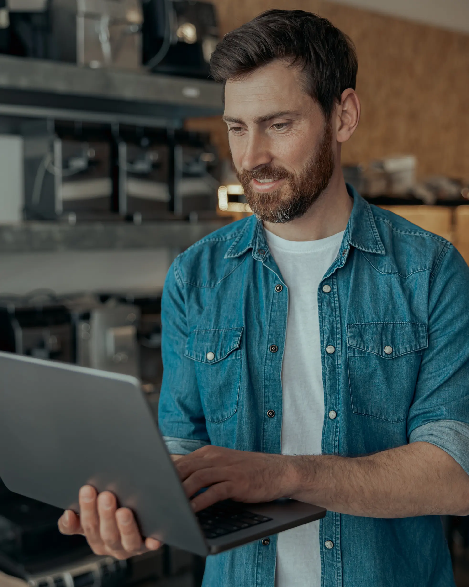 Mann mit Bart trägt ein blaues Jeanshemd und benutzt einen Laptop in einer Werkstatt oder Lagerraum.