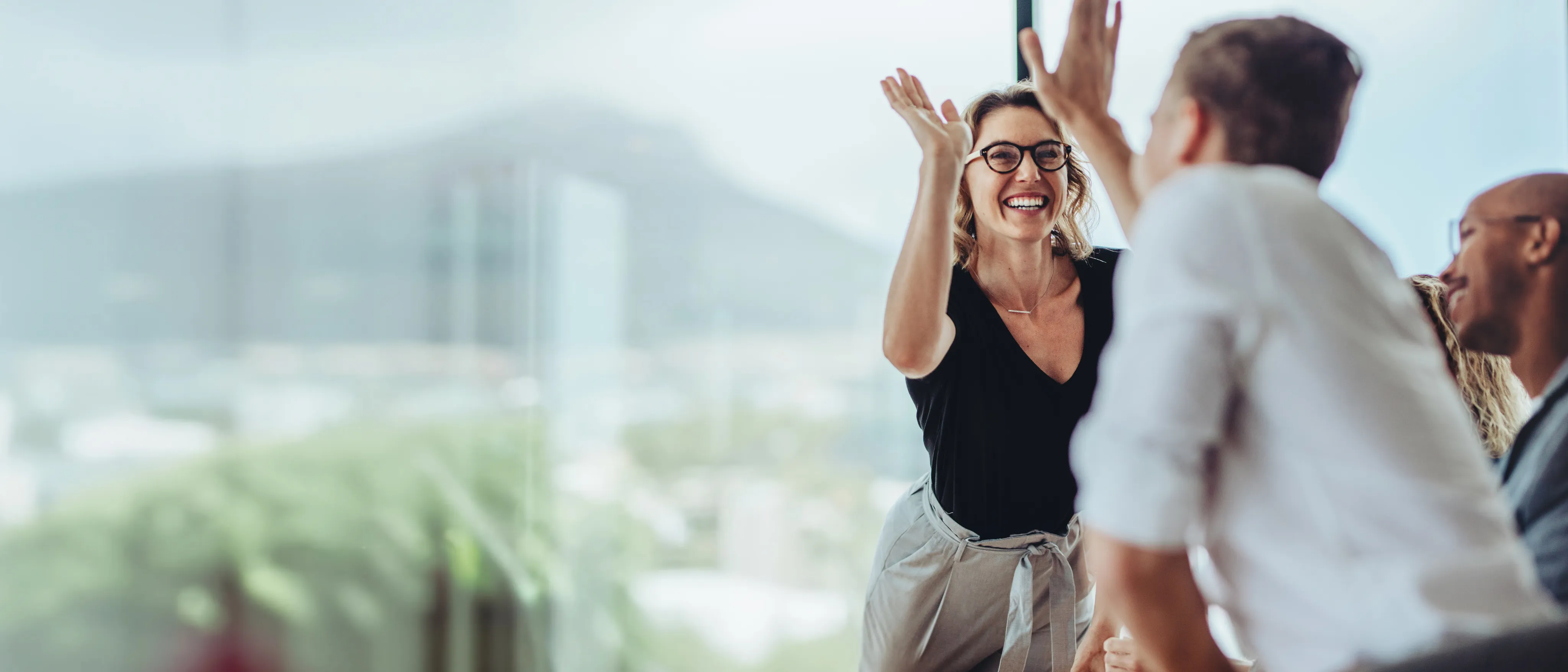 Lächelnde Frau mit Brille gibt einem Mann einen High-Five in einem modernen Büro mit Blick auf Berge.