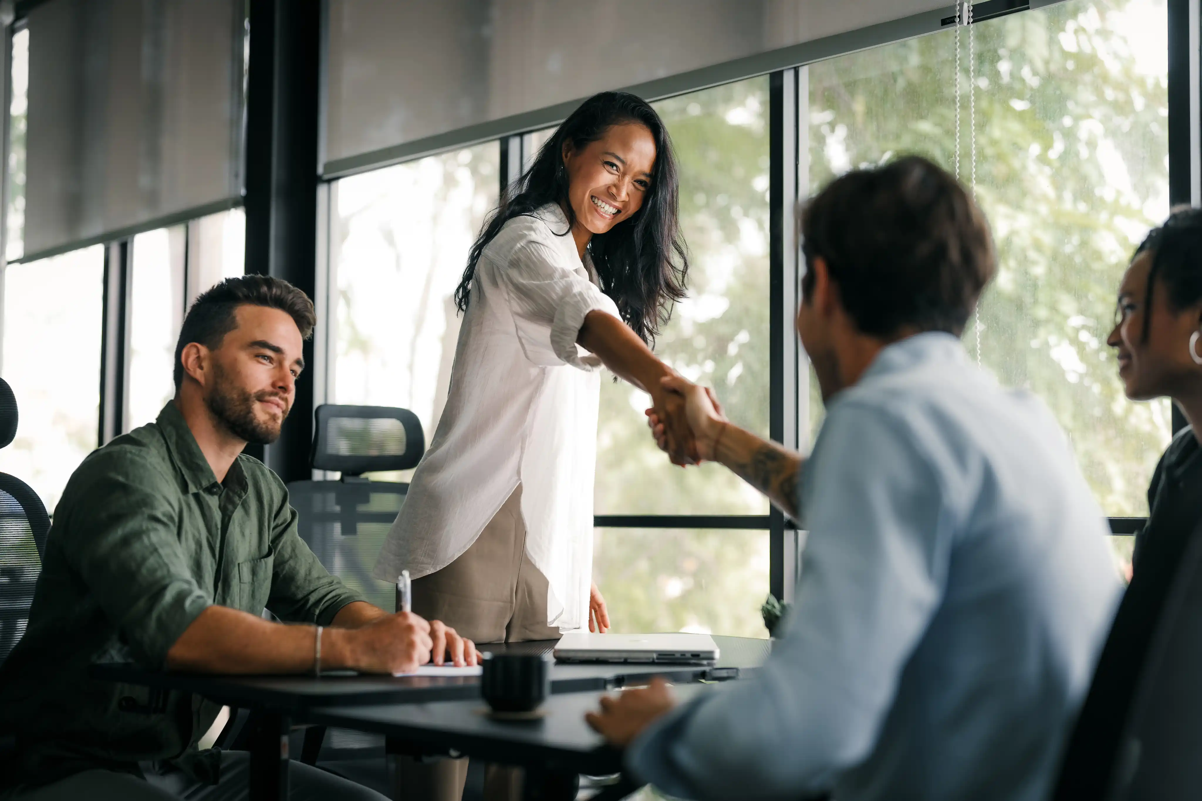 Frau und Mann geben sich in einem modernen Büro lächelnd die Hand, während zwei Kollegen am Tisch sitzen.