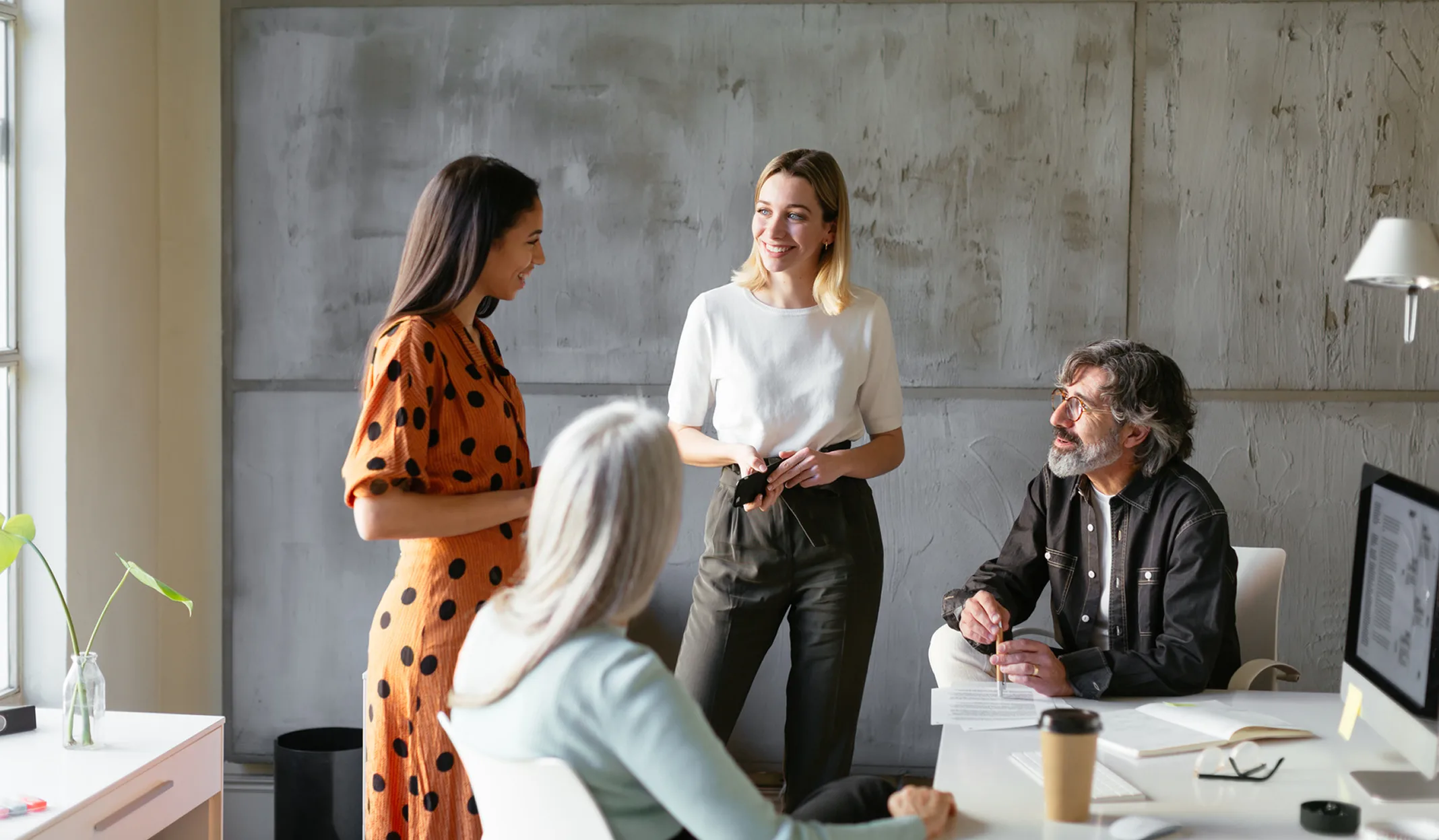 Vier Kollegen unterhalten sich in einem modernen Büro mit Betonwand, während sie an einem Schreibtisch mit Computerausrüstung sitzen.