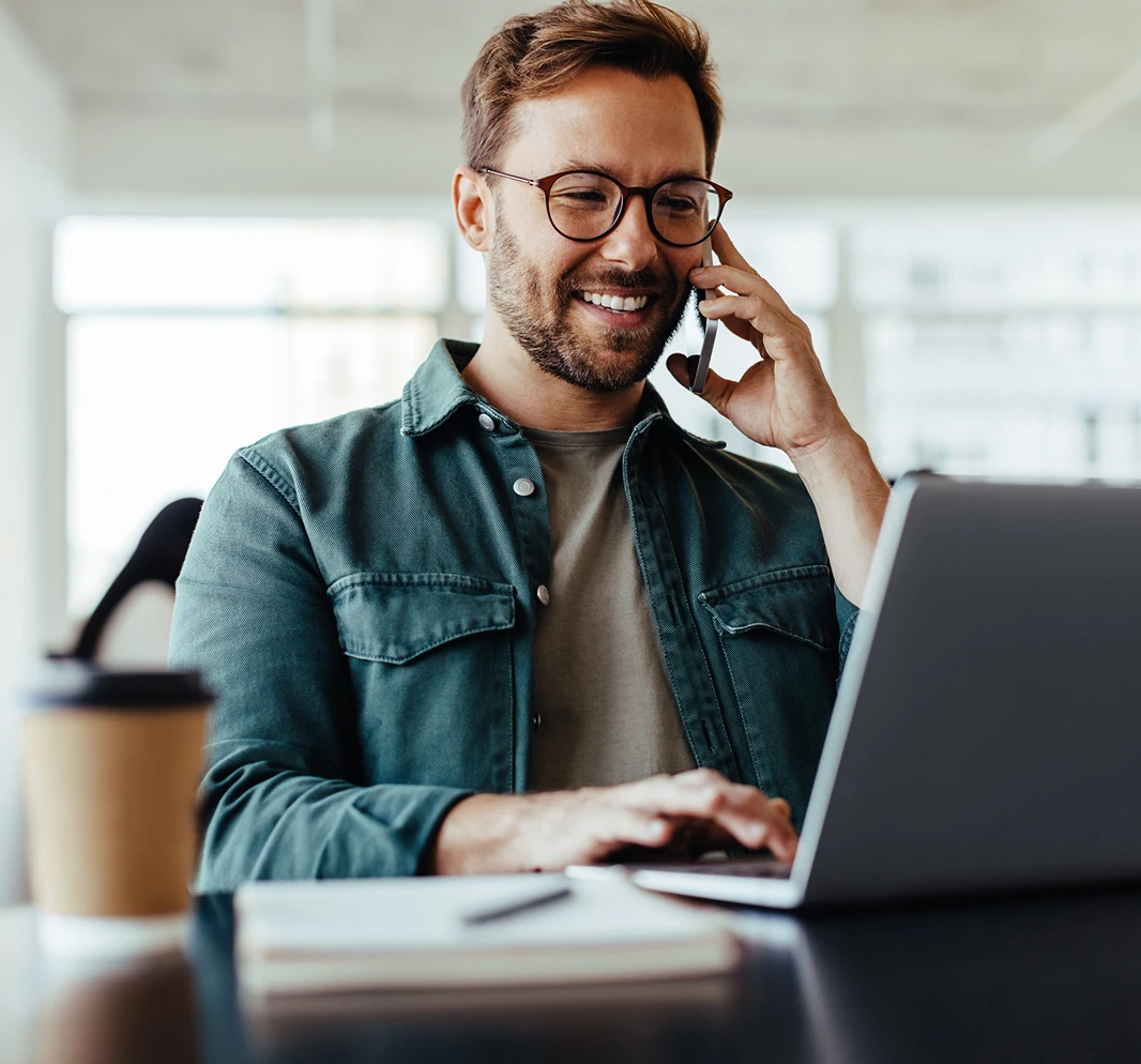 Mann mit Brille telefoniert lächelnd und tippt auf einem Laptop in einem hellen Büro.