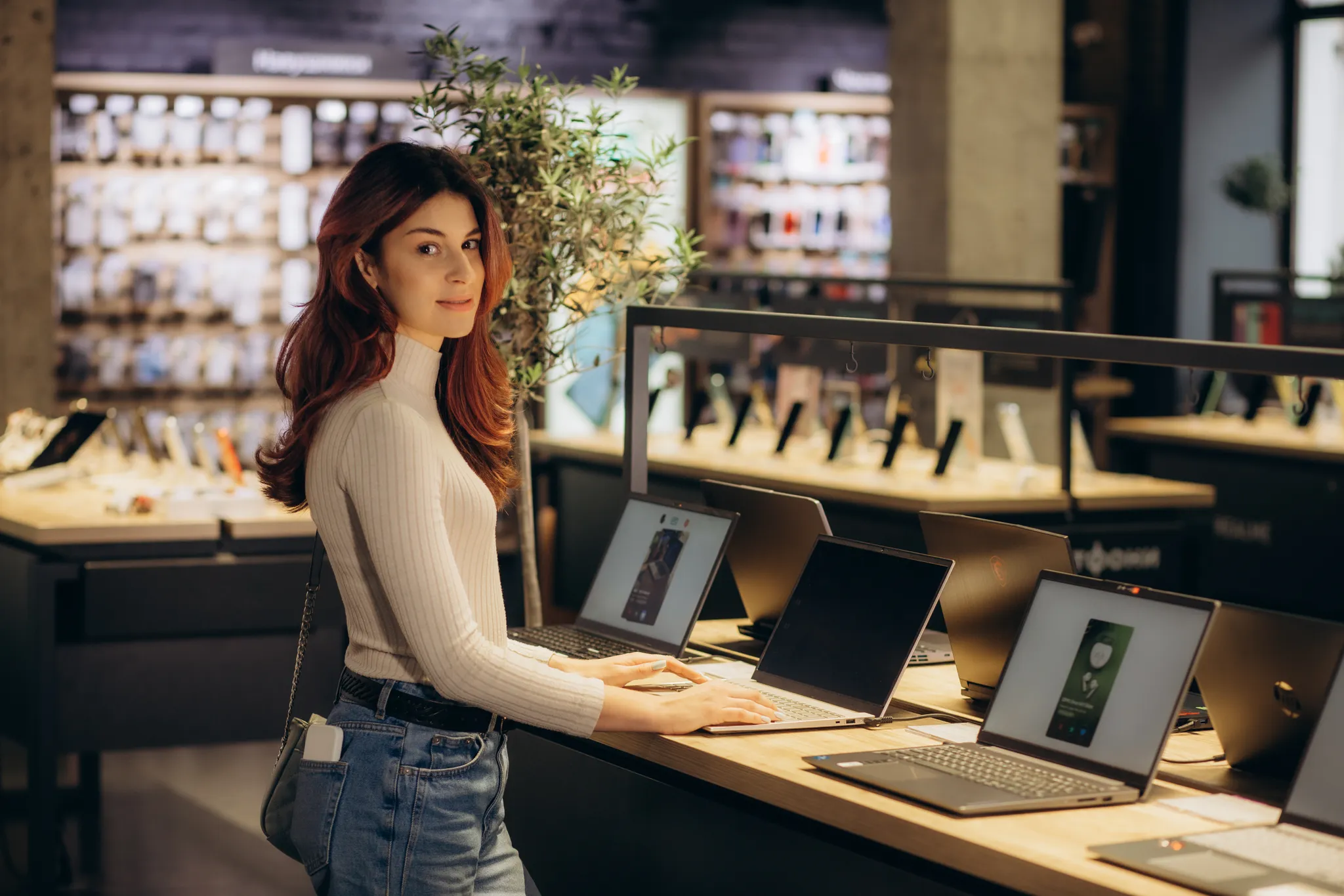 Eine Frau in einem weißen Rollkragenpullover steht an einem Tisch und testet Laptops in einem modernen Elektronikgeschäft.