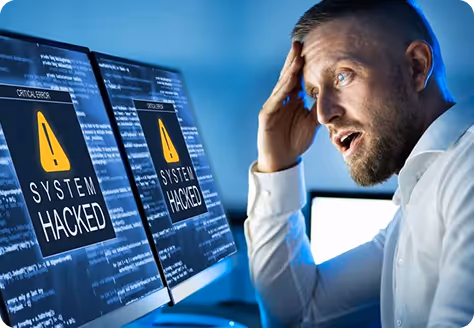 Man in white shirt looking shocked at dual computer monitors displaying 'System Hacked' warning messages.