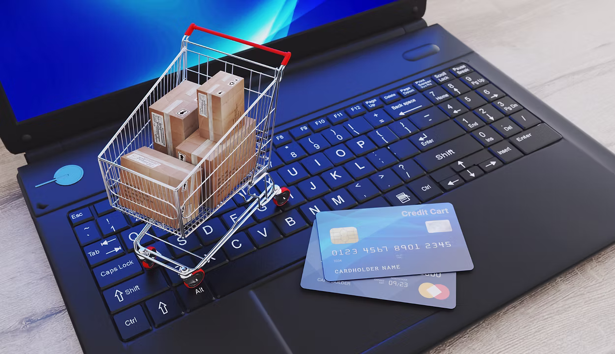 Miniature shopping cart filled with small boxes is placed on a laptop keyboard next to two credit cards.