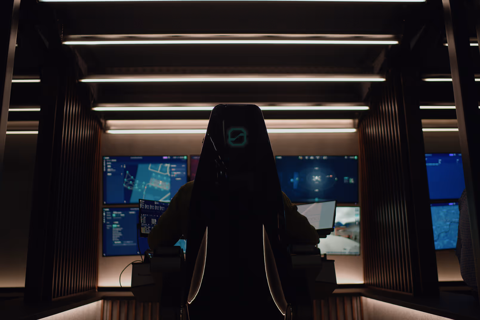 Person sitting in a high-back chair facing multiple illuminated computer screens in a dark, modern control room.