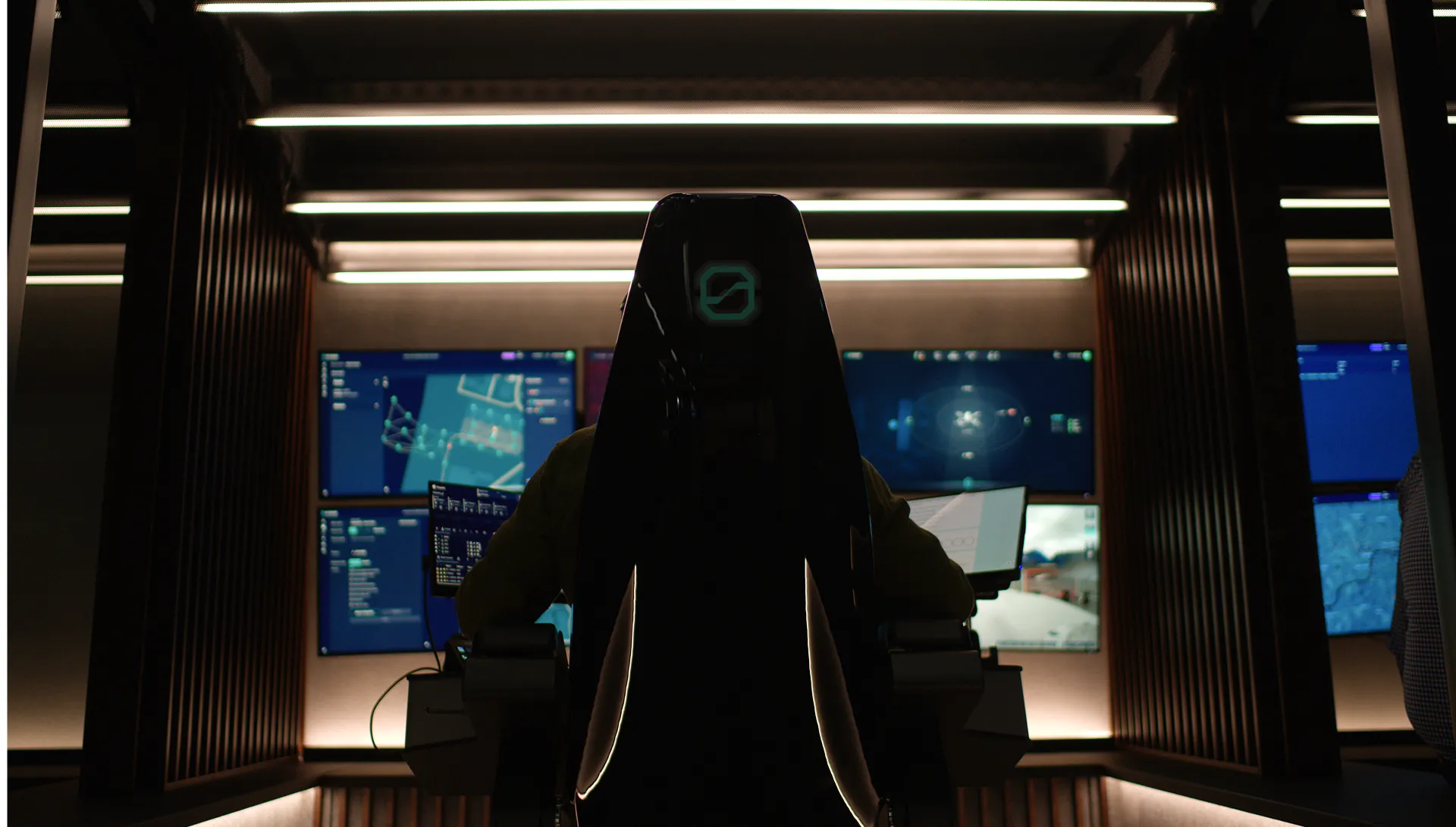 Person sitting in a dark control room facing multiple large screens displaying maps and data.