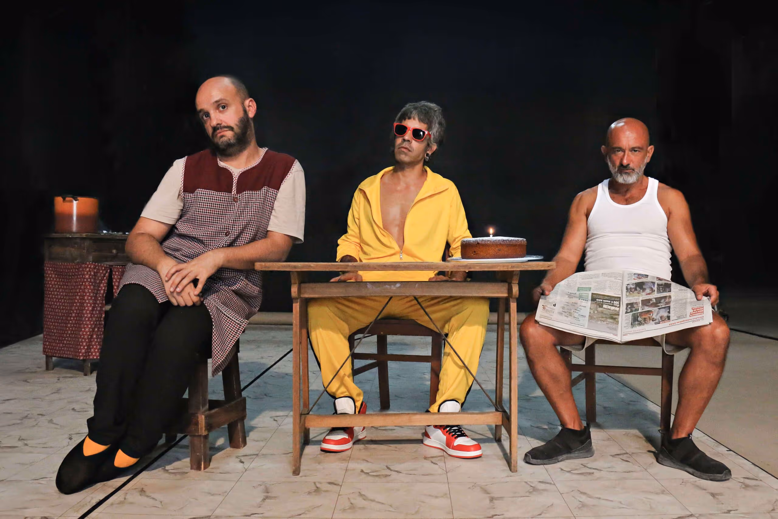 Three men sitting on chairs around a wooden table; the left man wears a checked apron, the middle man wears a yellow tracksuit and red sunglasses with a birthday cake on the table, and the right man wears a white tank top holding a newspaper.