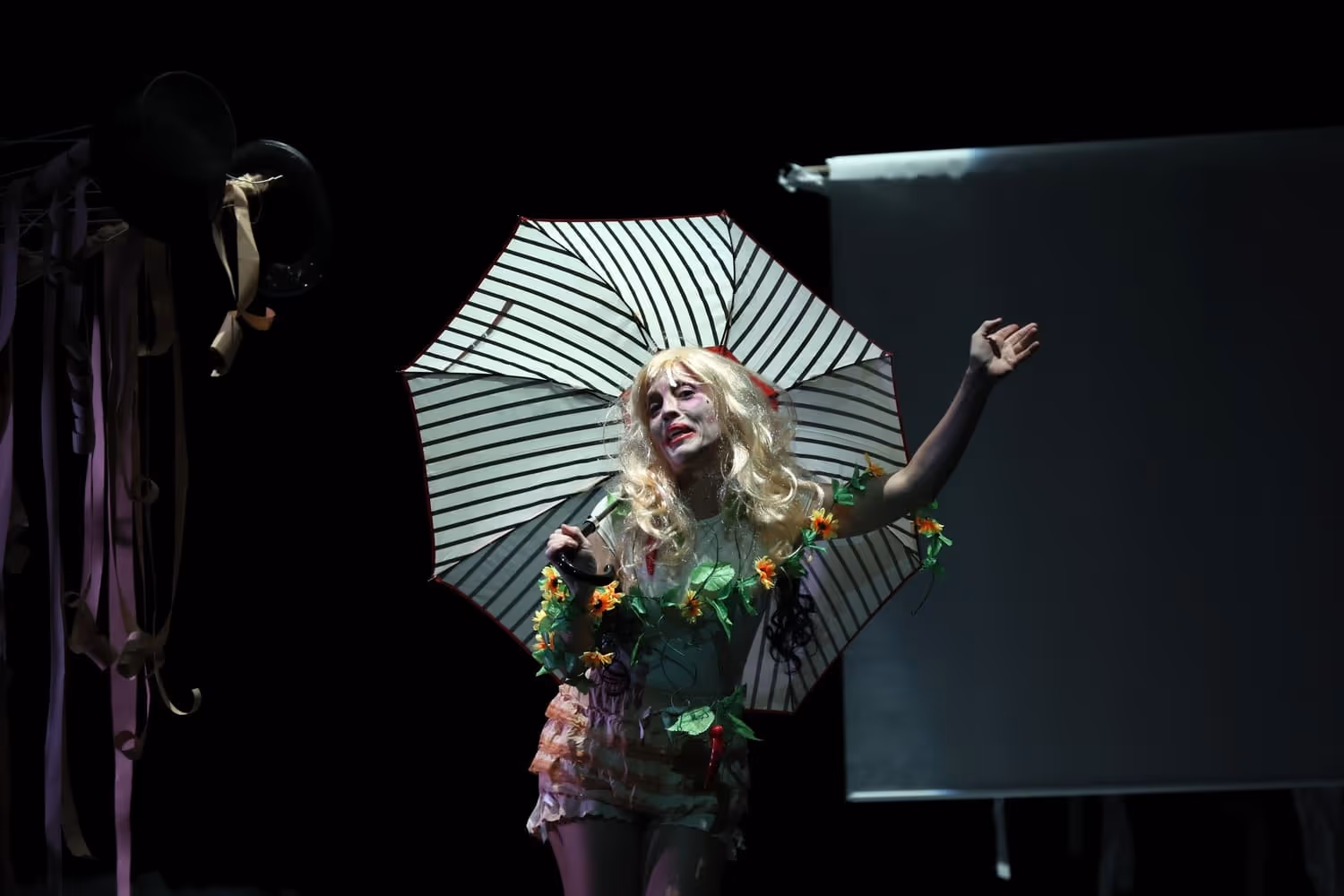 Ator vestido com peruca loira, maquiagem teatral e roupa decorada com flores, segurando um guarda-chuva listrado aberto num palco escuro.
