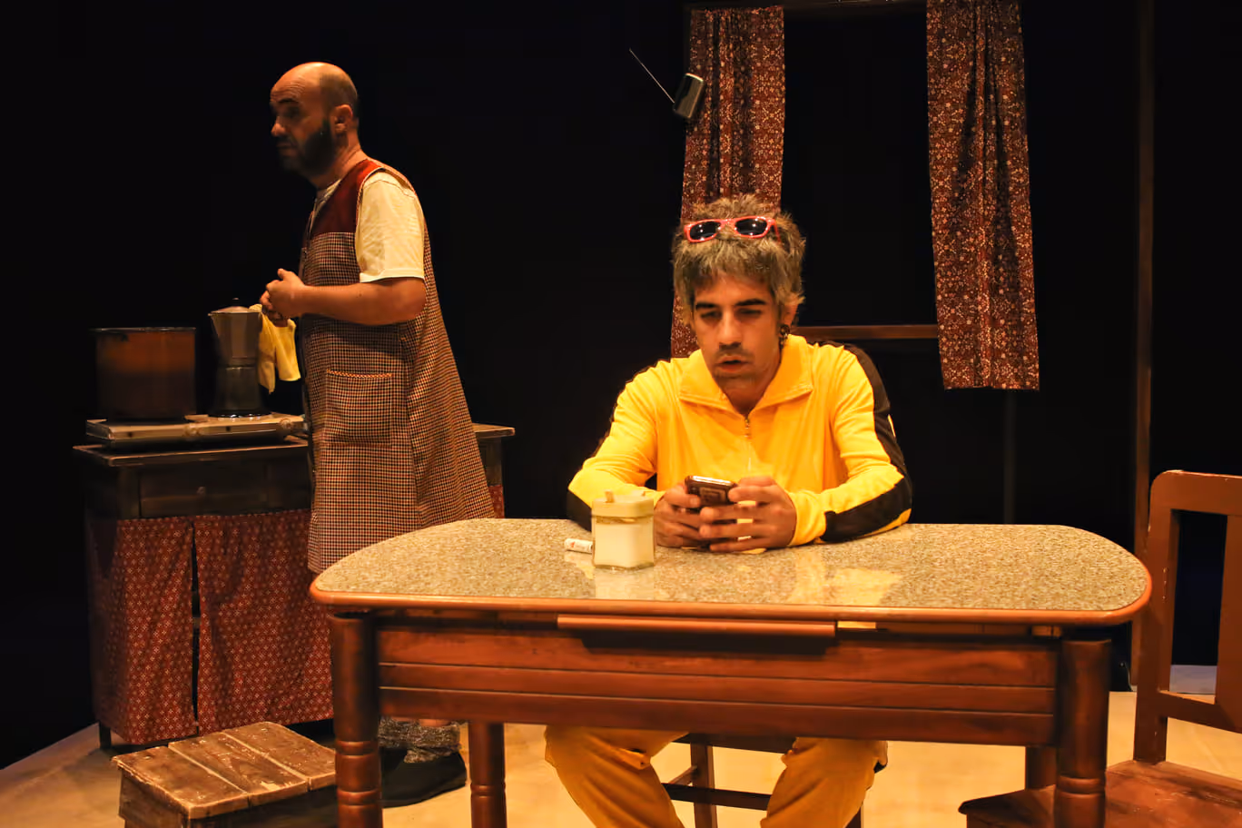 Cena de teatro com homem de colete a limpar uma cafeteira e jovem sentado à mesa usando fato amarelo e a olhar para um telemóvel.