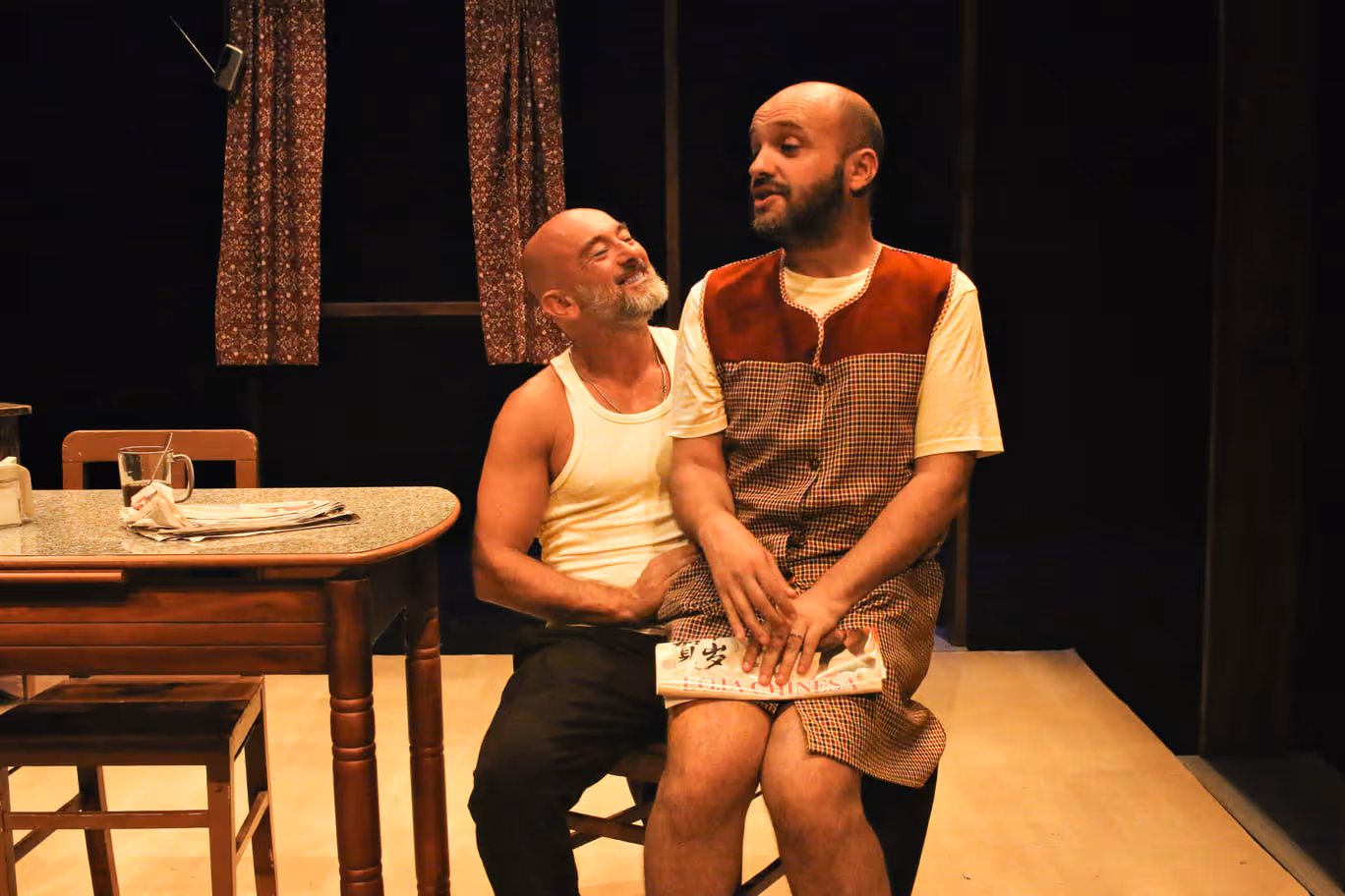 Dois homens carecas sentados juntos num cenário teatral com uma mesa e cortinas ao fundo.