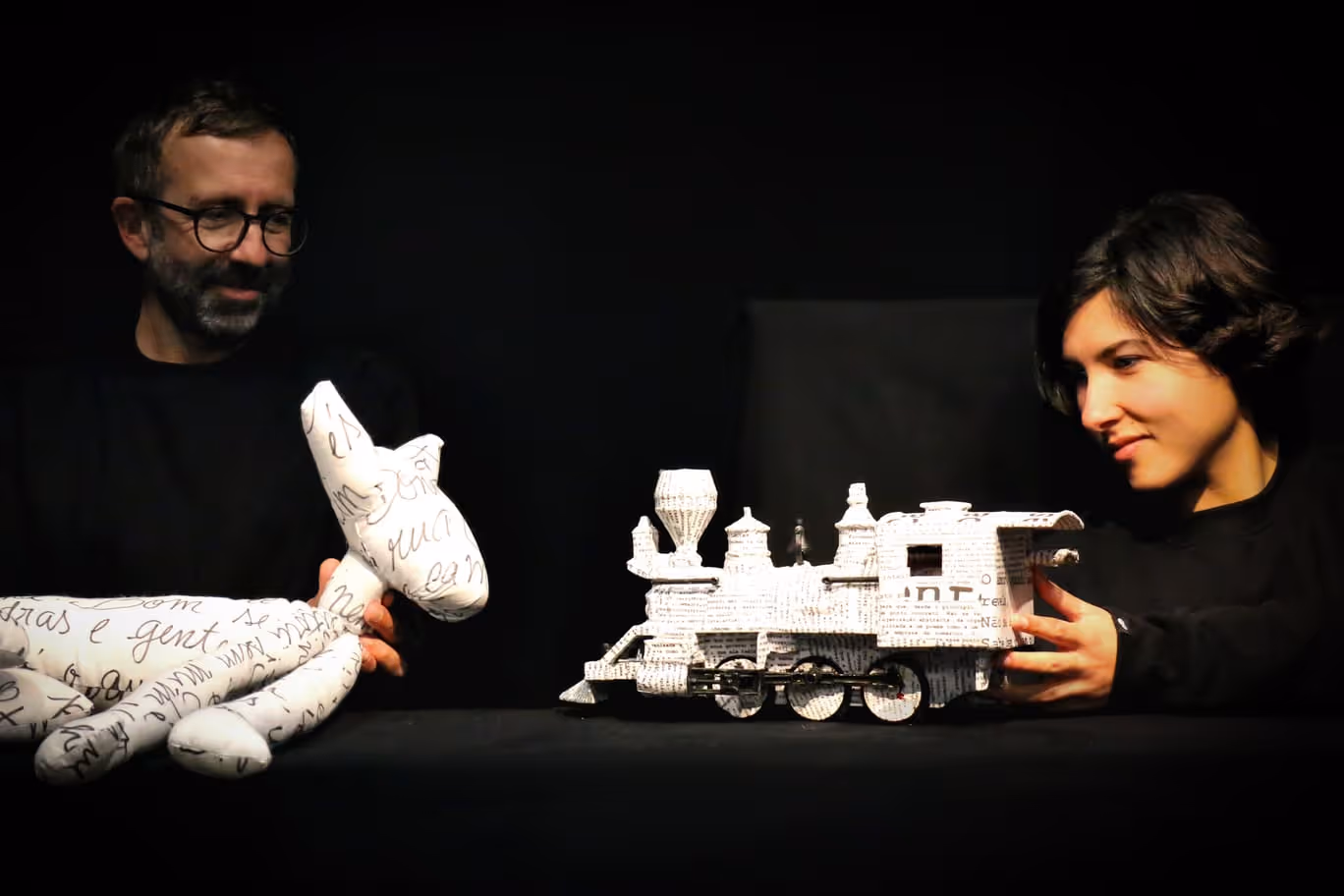 Um homem e uma mulher vestidos de preto seguram esculturas de papel branco representando um cão e um comboio sobre uma superfície preta.